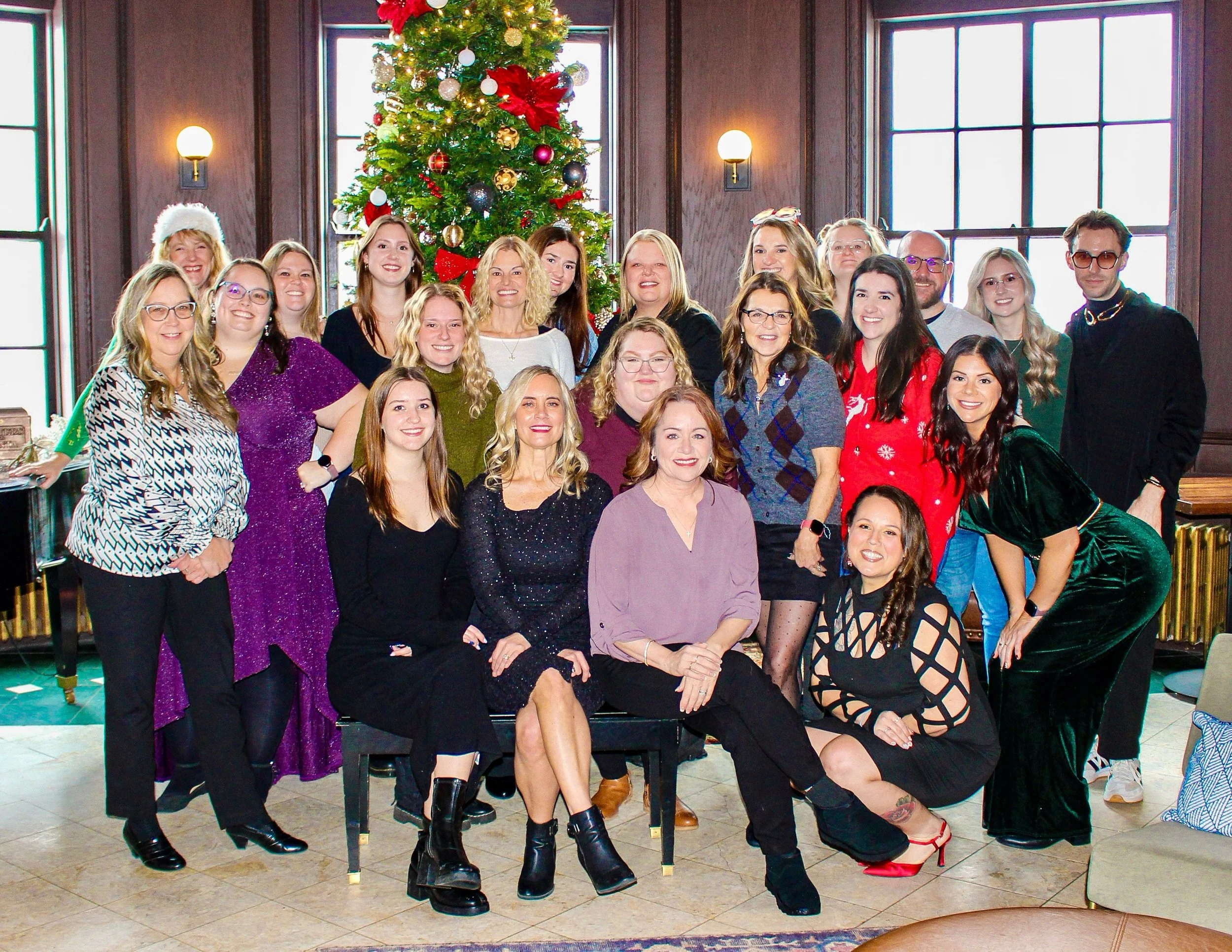A group of women and men gathered together around a decorated Christmas tree, smiling for the camera. They are in a room with large windows and wooden paneling, wearing festive clothing and accessories.