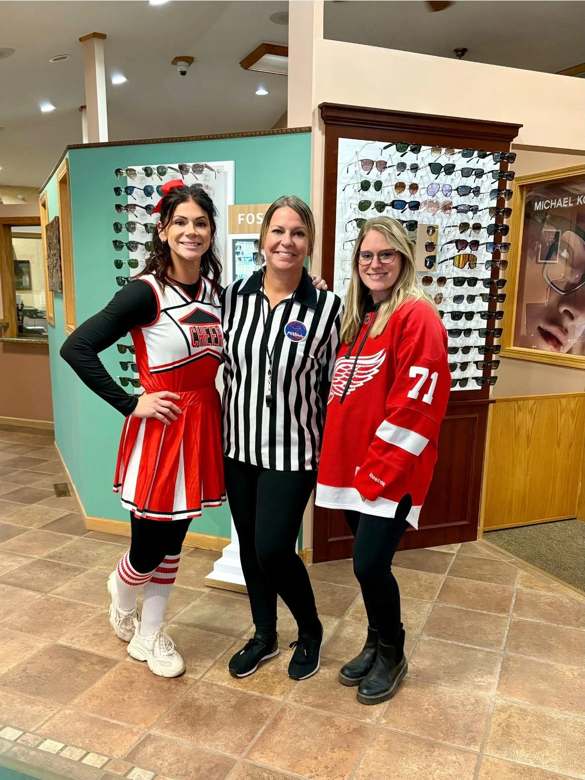 Three women standing indoors, one dressed as a cheerleader, another as a hockey player, and one in a referee outfit, smiling in front of sunglasses display.