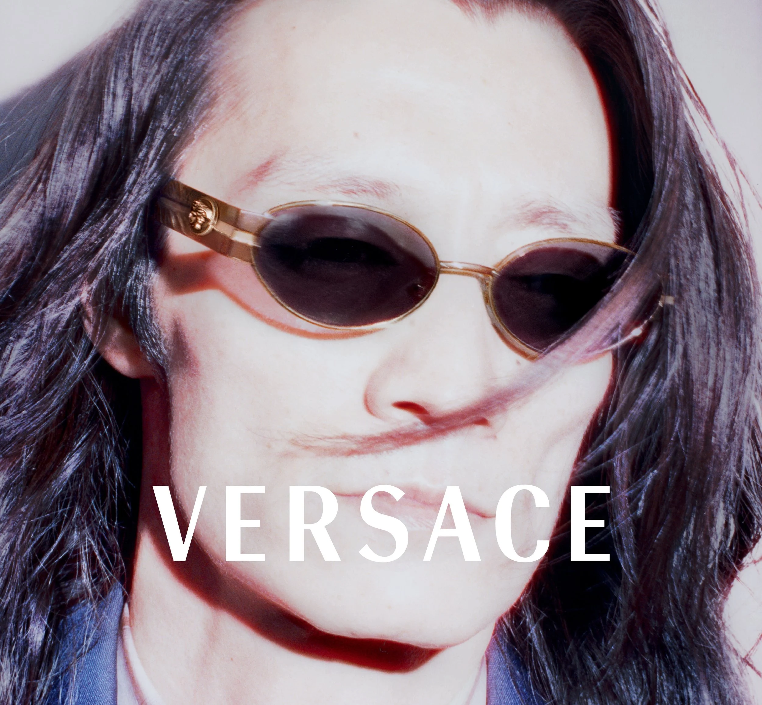 Close-up of a person's face with long dark hair, wearing dark sunglasses with gold embellishments on the temples, and large white text overlay reading "VERSACE."