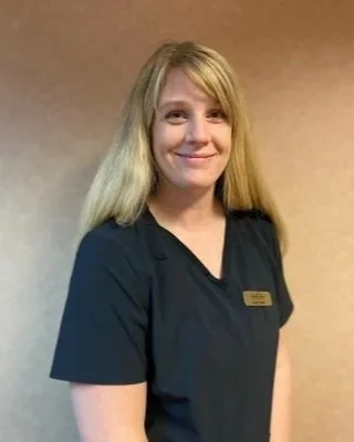 A woman with long blonde hair wearing a dark uniform and a name badge, smiling at the camera, standing against a plain background.