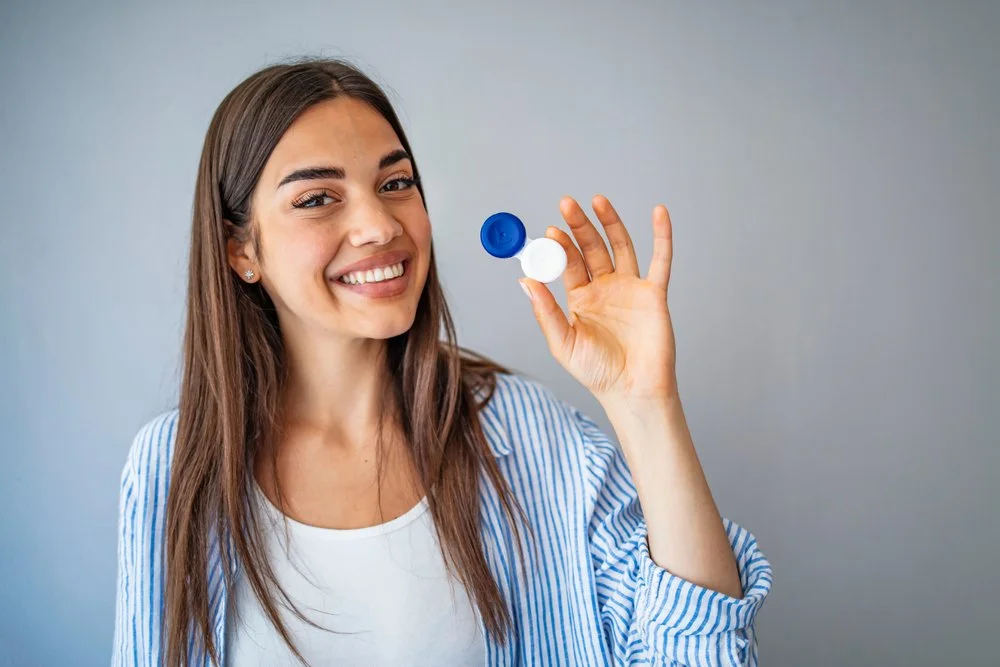 Smiling woman holding a contact lens case with the lid open, showing the lenses inside.