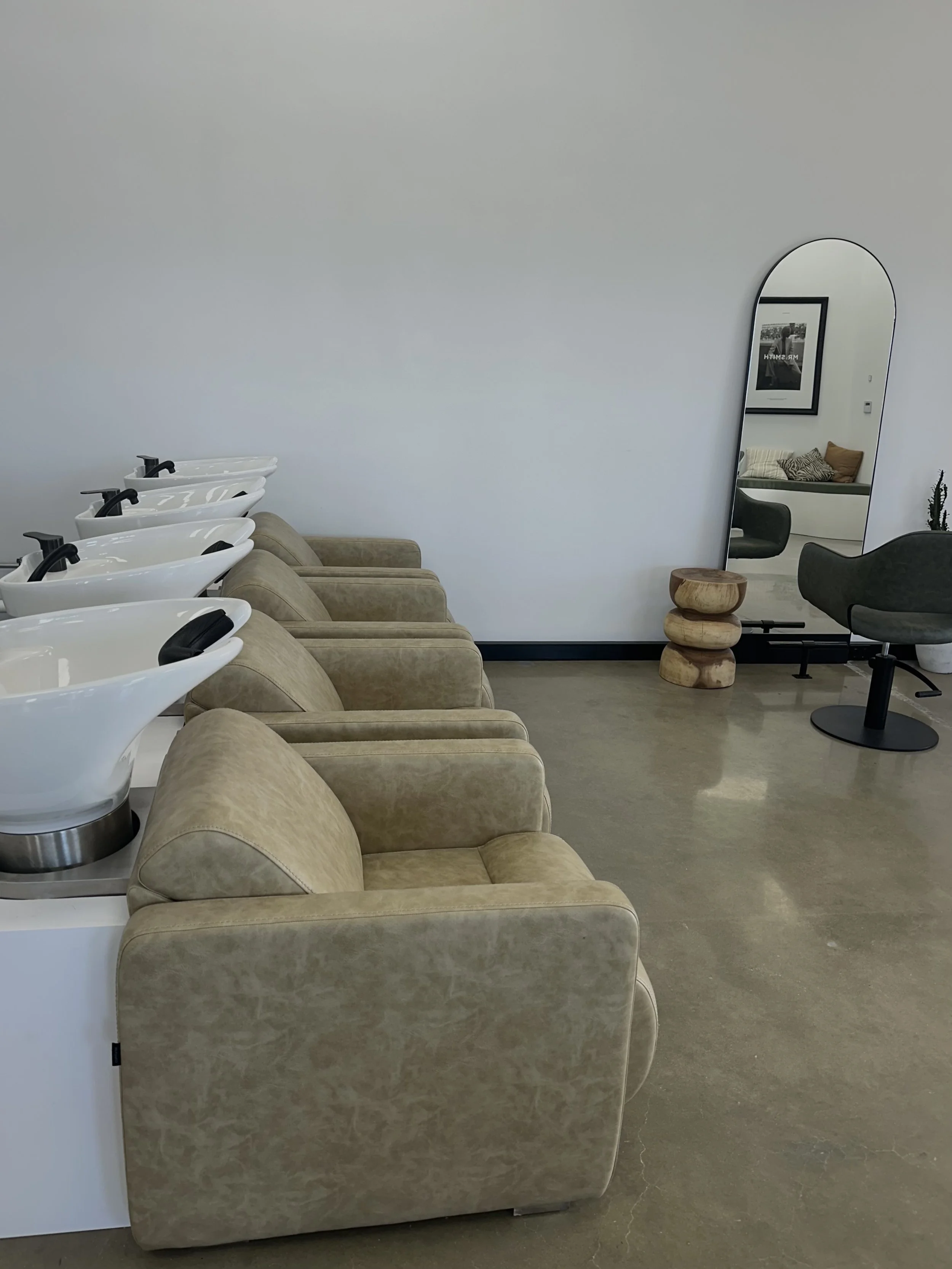 Salon with four beige chairs and three white hair washing sinks lined up along a white wall, a tall curved mirror, a small wooden stool, and a black salon chair with a gray cushion.