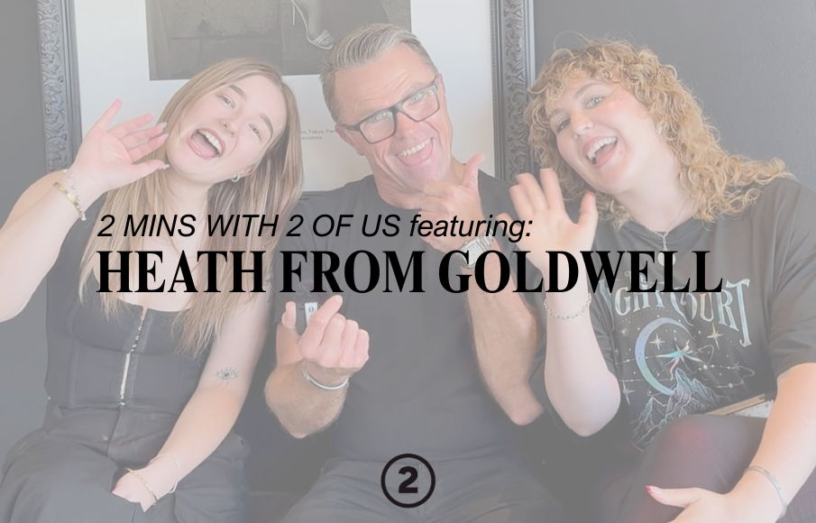 Three people sitting together, two women and a man, smiling and waving at the camera. They are indoors, with framed artwork on the wall behind them.