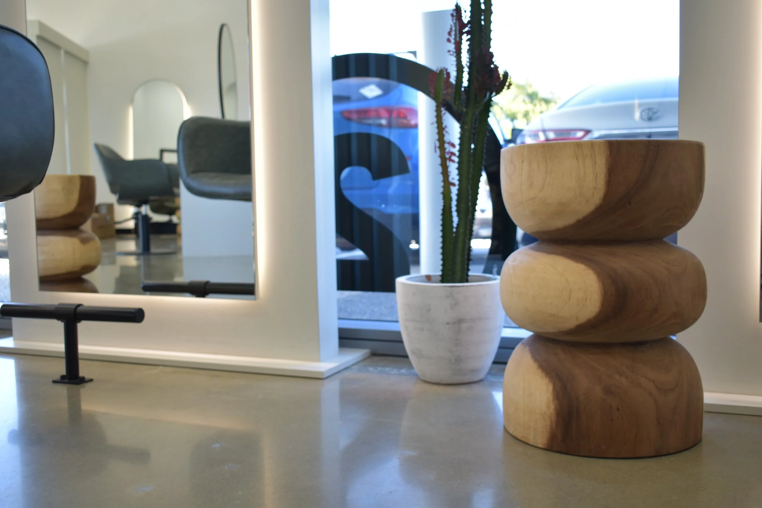 Interior view of a modern space with a stacked wooden sculpture, a potted cactus plant, and a mirror reflecting chairs and furniture.