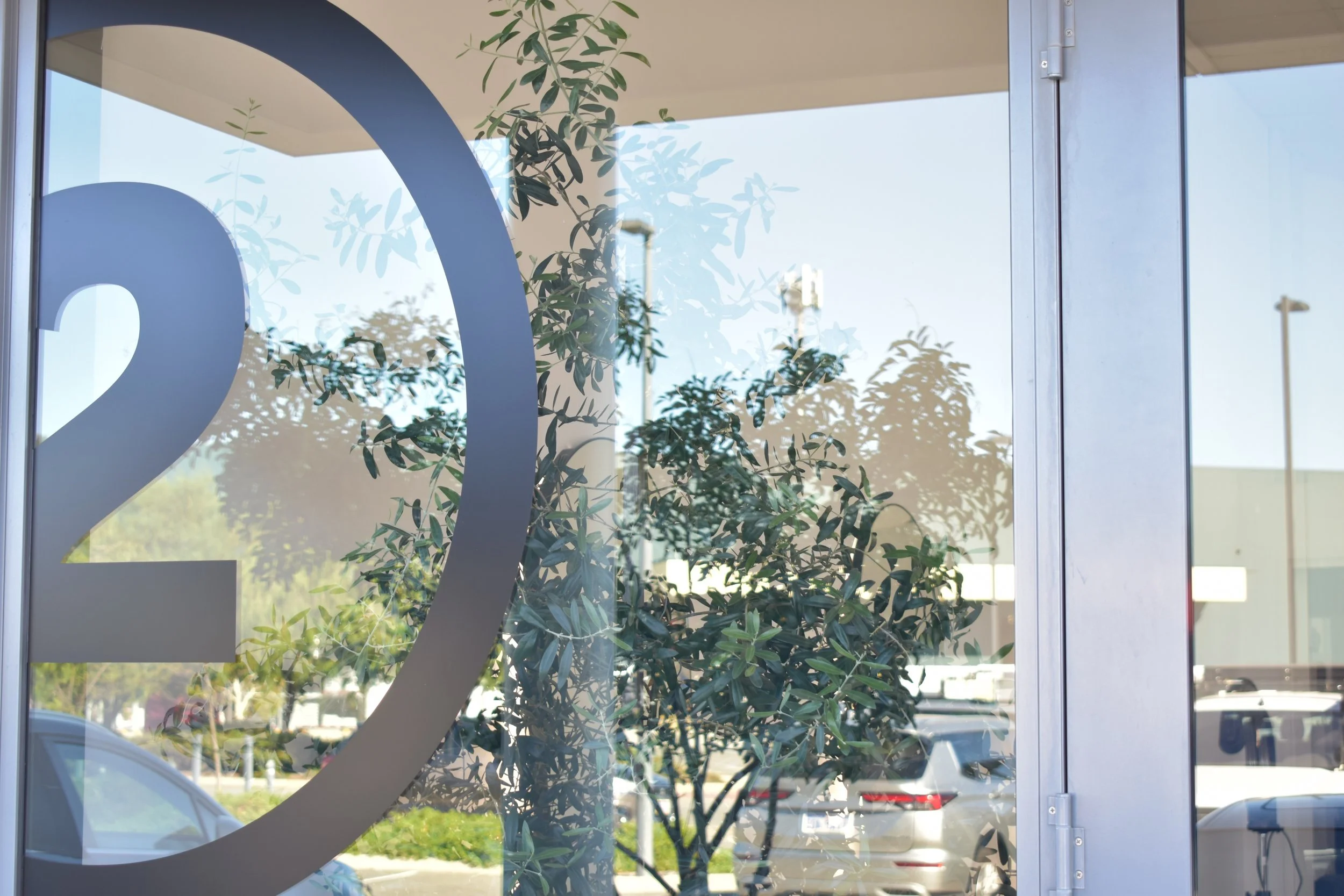 View through a glass window with a large number 2 (Salons logo and signature signage) printed on it, inside olive tree as decoration and parked cars outside.