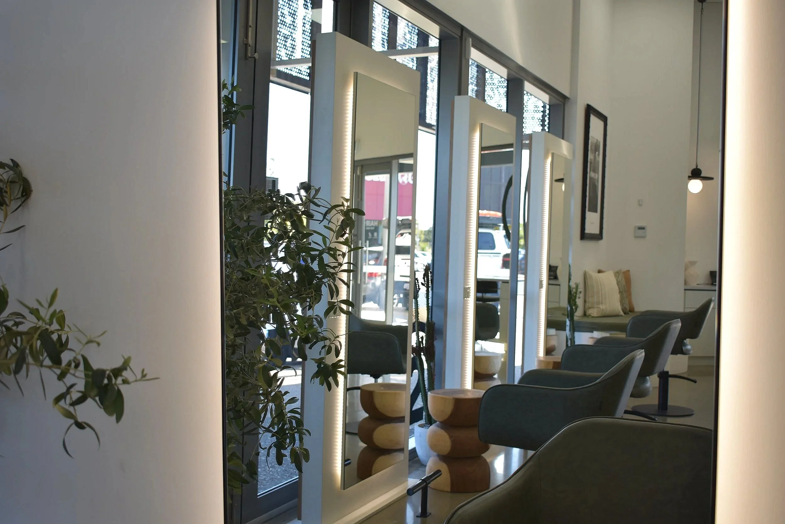 Interior of a modern salon with styling chairs, mirrors with lighting, and decorative wooden stools, inside a room with large windows letting in natural light.