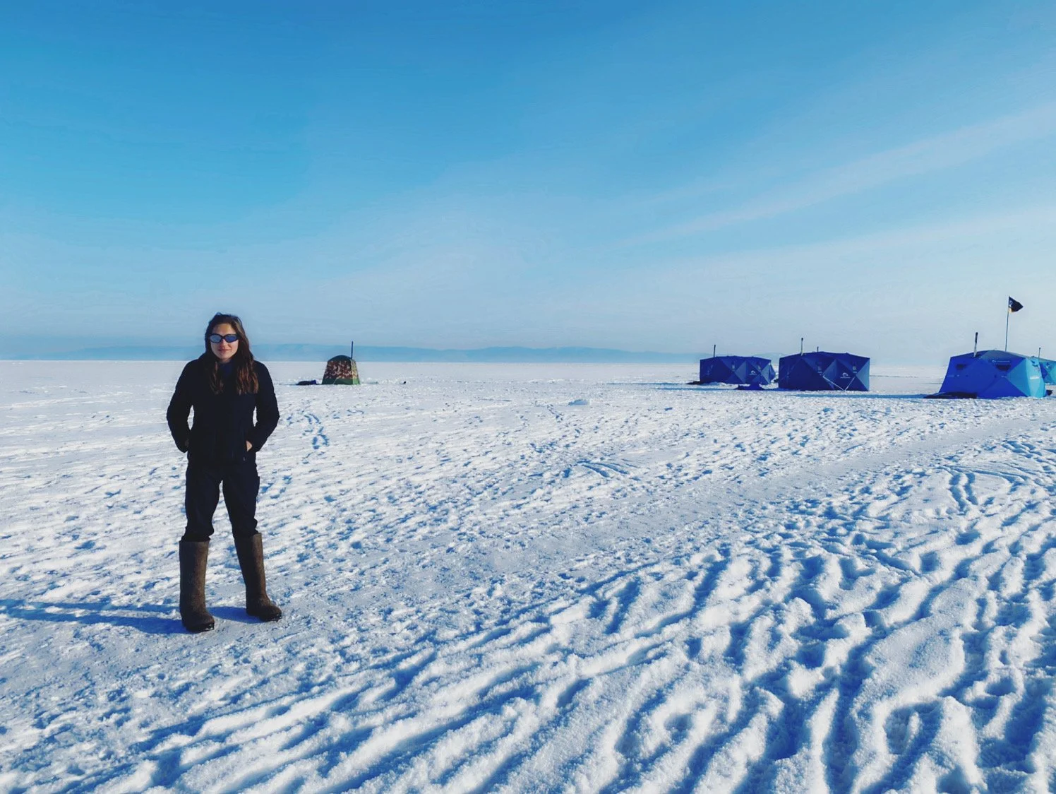 A woman in black winter clothing and tall boots standing on a snow-covered landscape with a clear blue sky, several blue tents, and distant mountains.