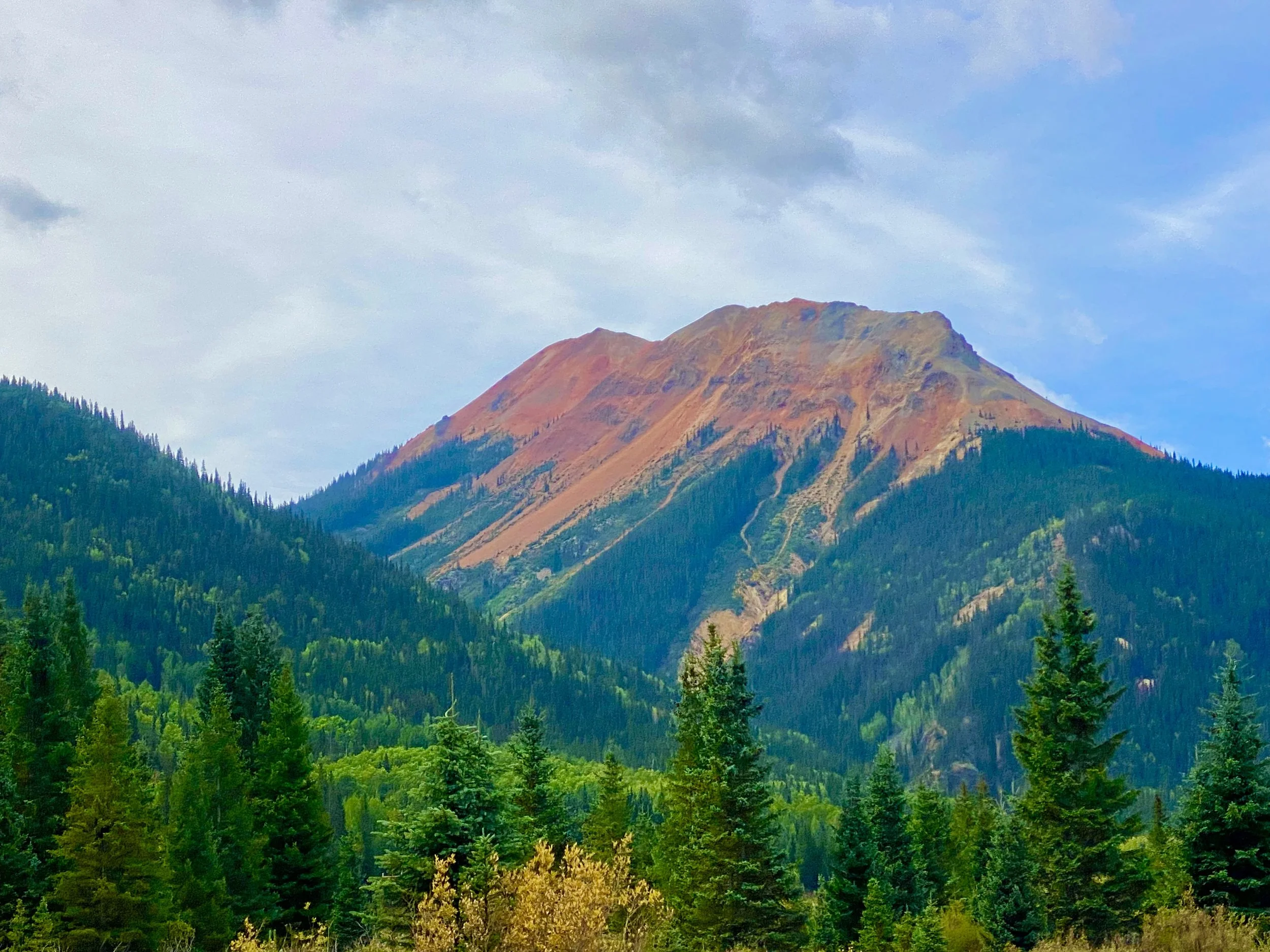 A mountain with a reddish-brown peak surrounded by green forest under a partly cloudy sky.