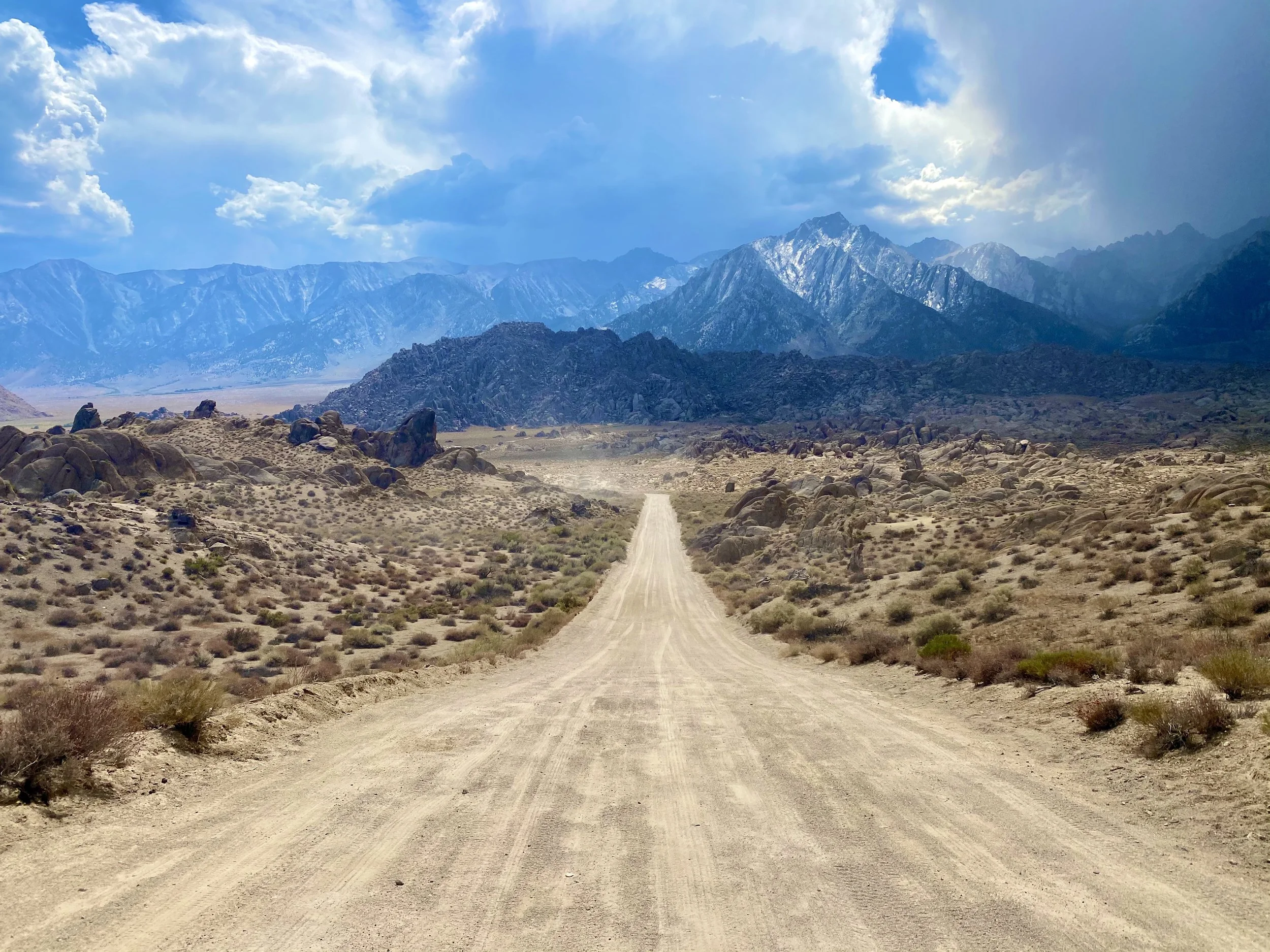 A dirt road runs through a desert landscape with scattered bushes and rocks, leading towards distant mountains with some snow-capped peaks under a partly cloudy sky.