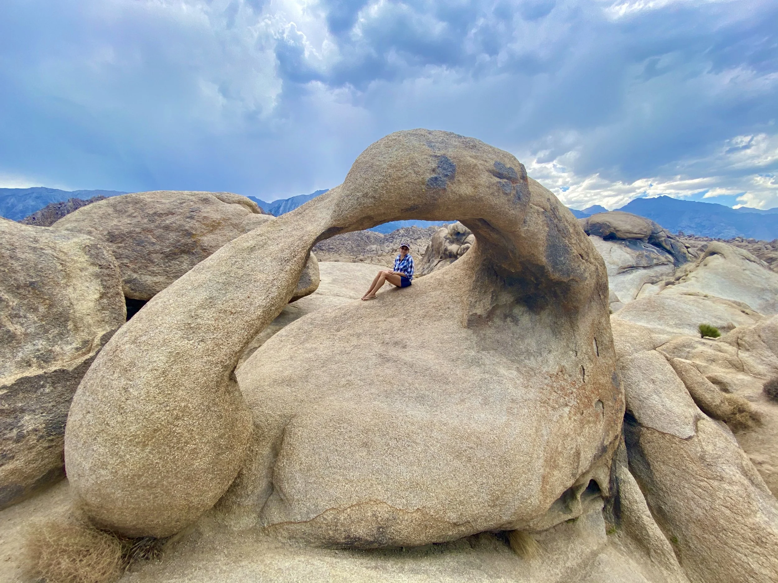 Person sitting inside a natural rock formation with a hole in the middle, in a desert landscape with mountains and cloudy sky in the background.