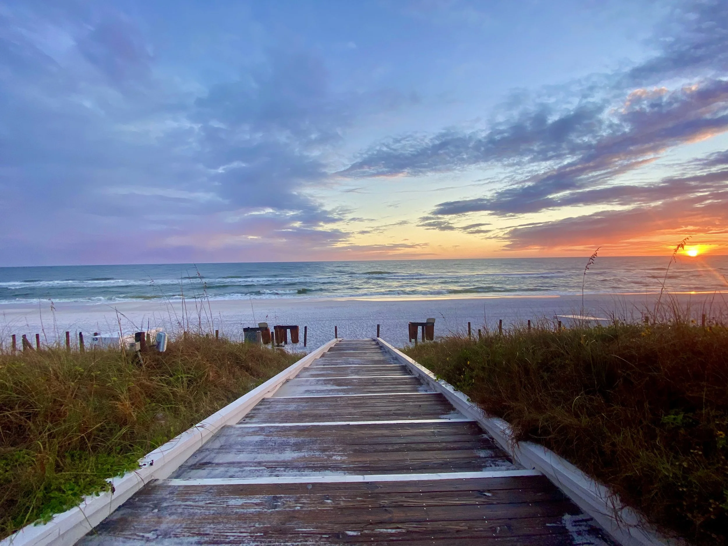 Wooden walkway leading to a beach with ocean and sunset, with cloudy sky and grassy dunes on sides.
