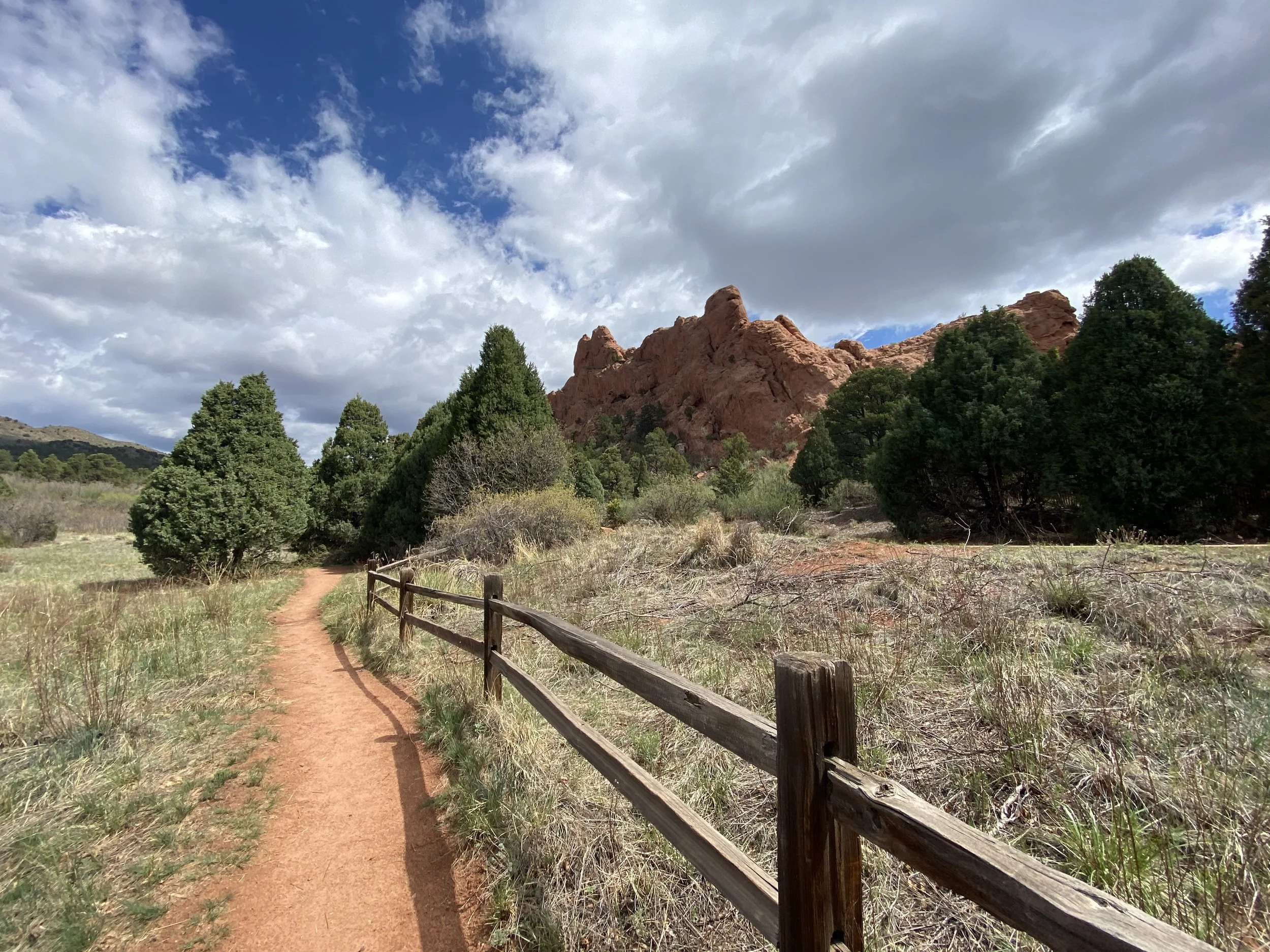 A dirt hiking trail with a wooden fence on the side, surrounded by green trees and red rock formations under a partly cloudy sky.