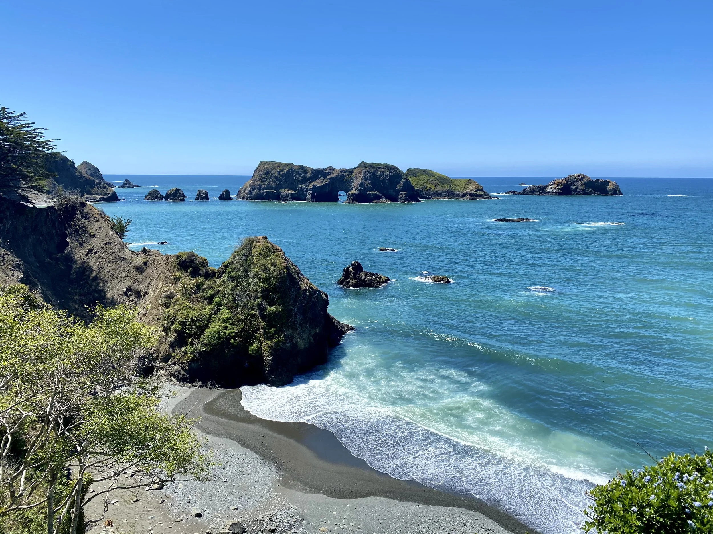 Scenic view of a rocky coastline with green hills, a small beach, and blue ocean under a clear sky.