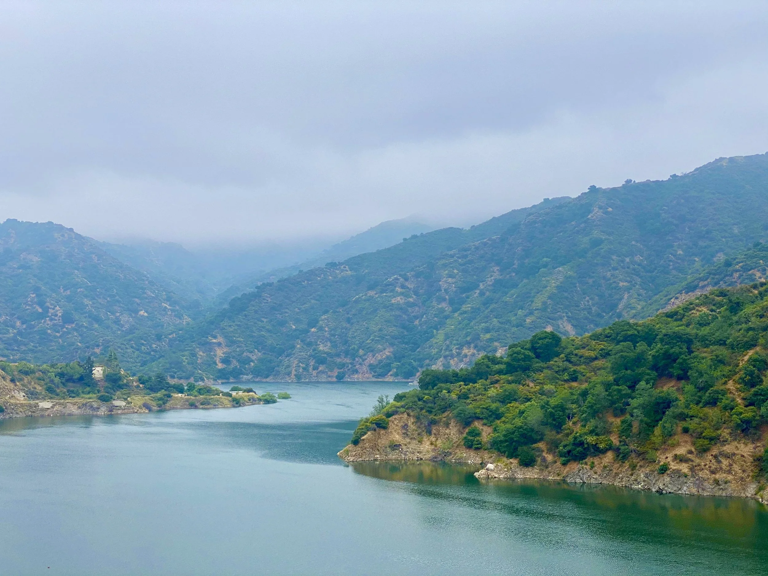 A scenic view of a river winding through green hills and mountains with a cloudy, overcast sky.