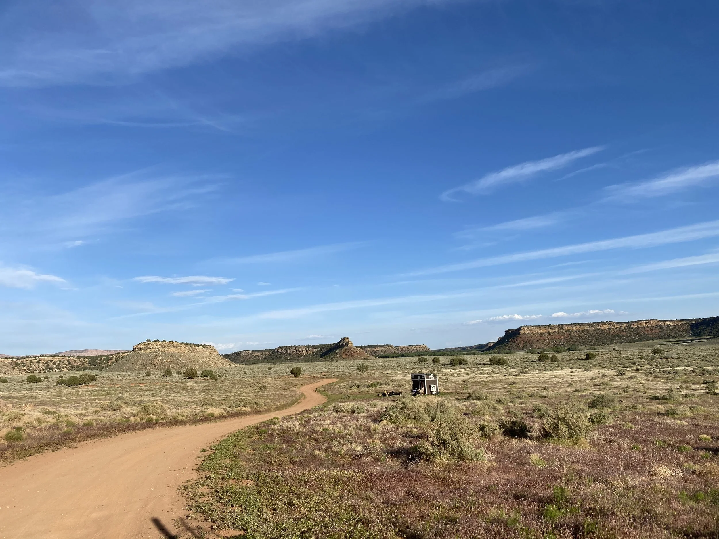 Desert landscape with a winding dirt road, sparse vegetation, flat-topped mesas in the background, and a clear blue sky with a few clouds.