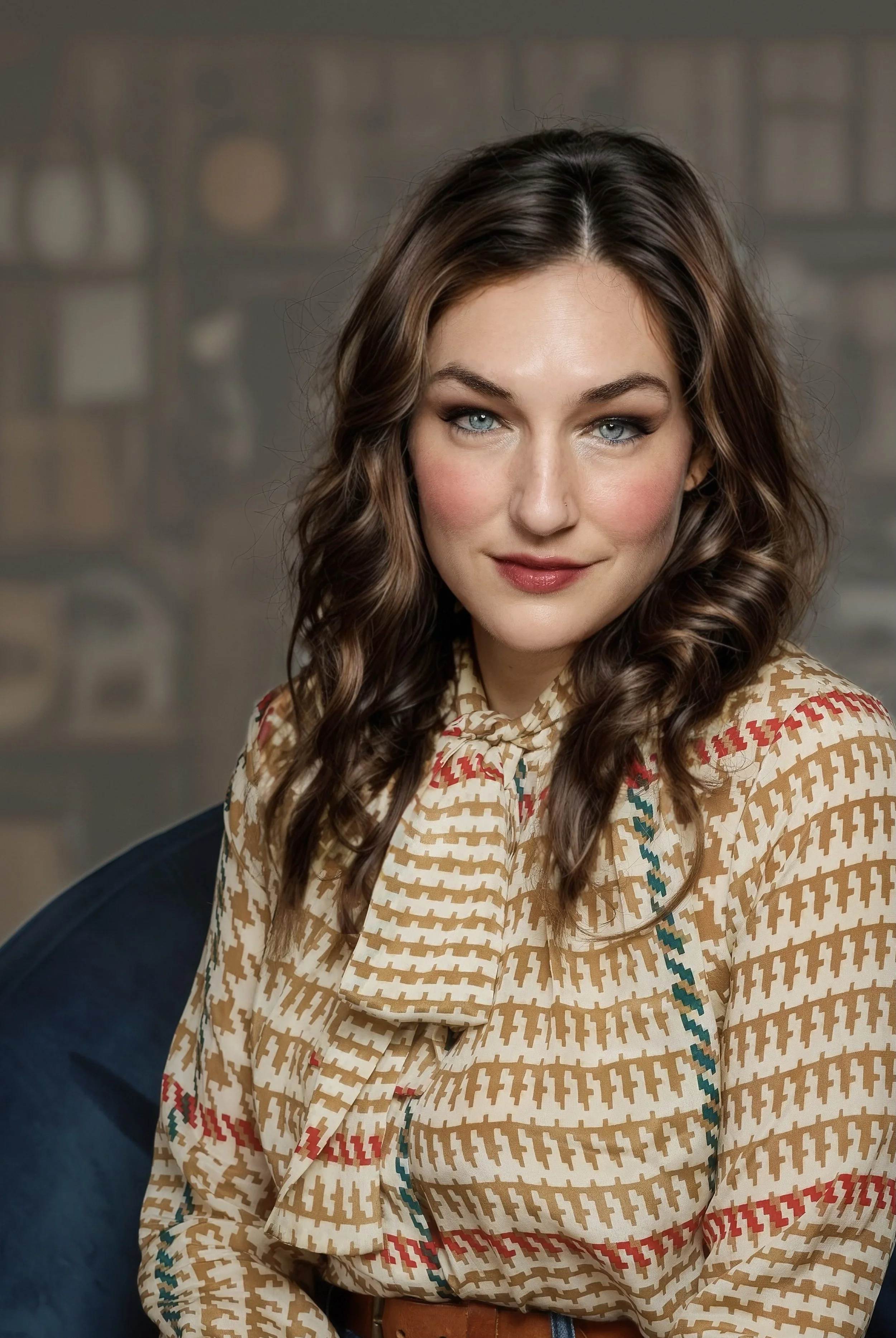 A woman with wavy brown hair and blue eyes, wearing a beige patterned blouse with a bow tie at the collar, sitting on a blue chair with a blurred bookcase background.