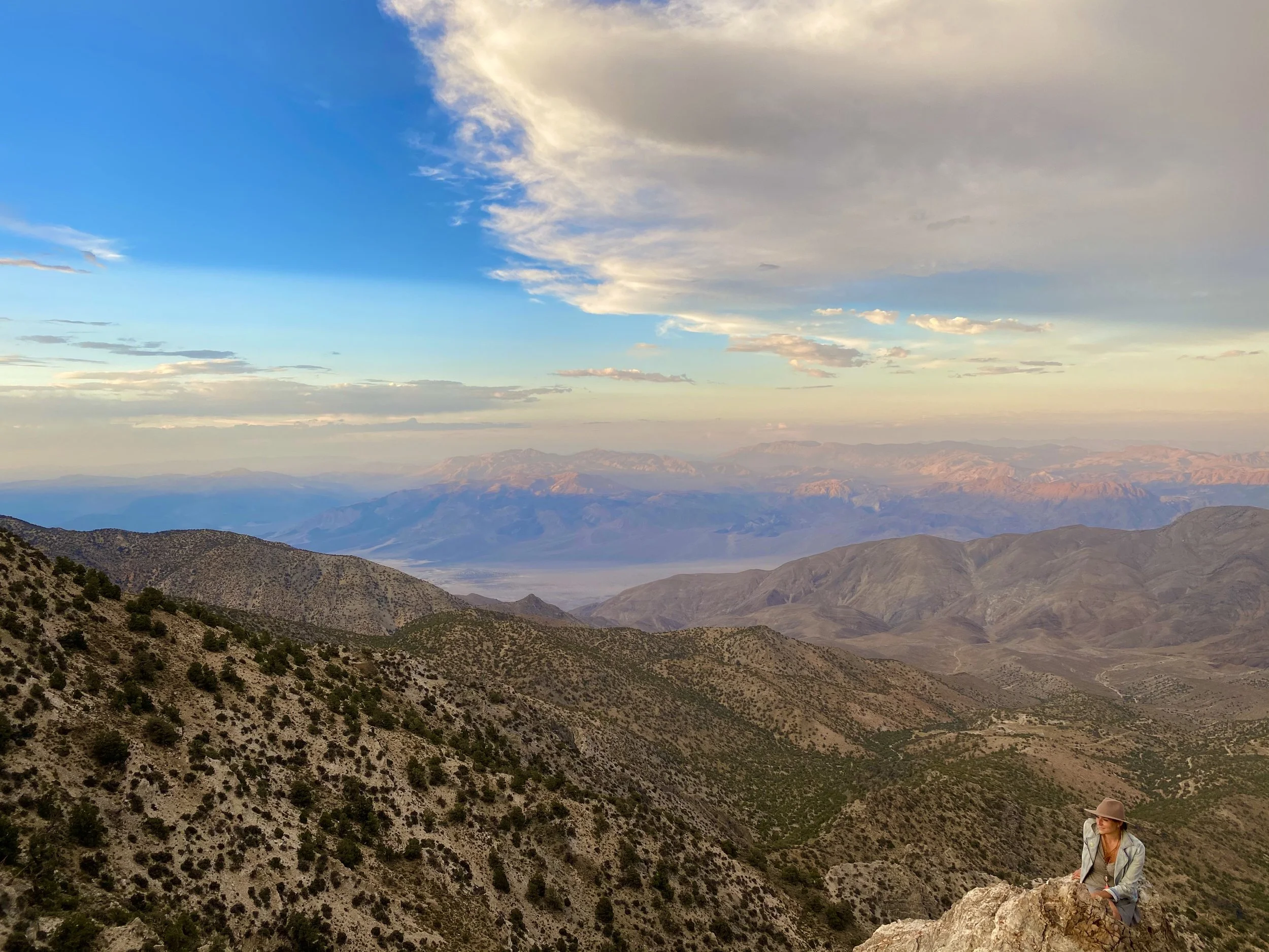 A woman in a hat and light jacket sitting on a rock on a mountain, overlooking a vast landscape with rolling hills, distant mountains, and a partly cloudy sky.