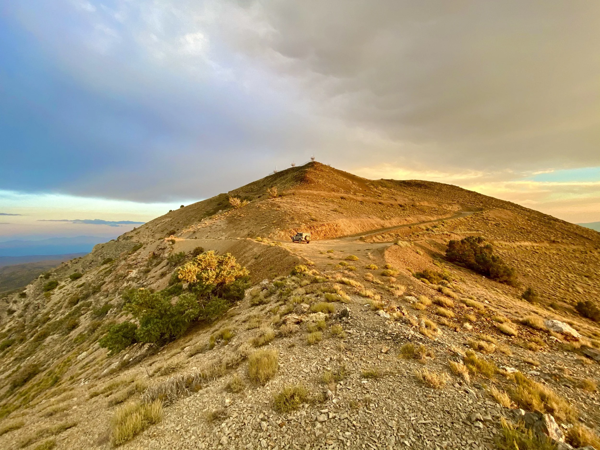 A rugged mountain trail with a vehicle parked along the side, overlooking a vast landscape under a cloudy sky.