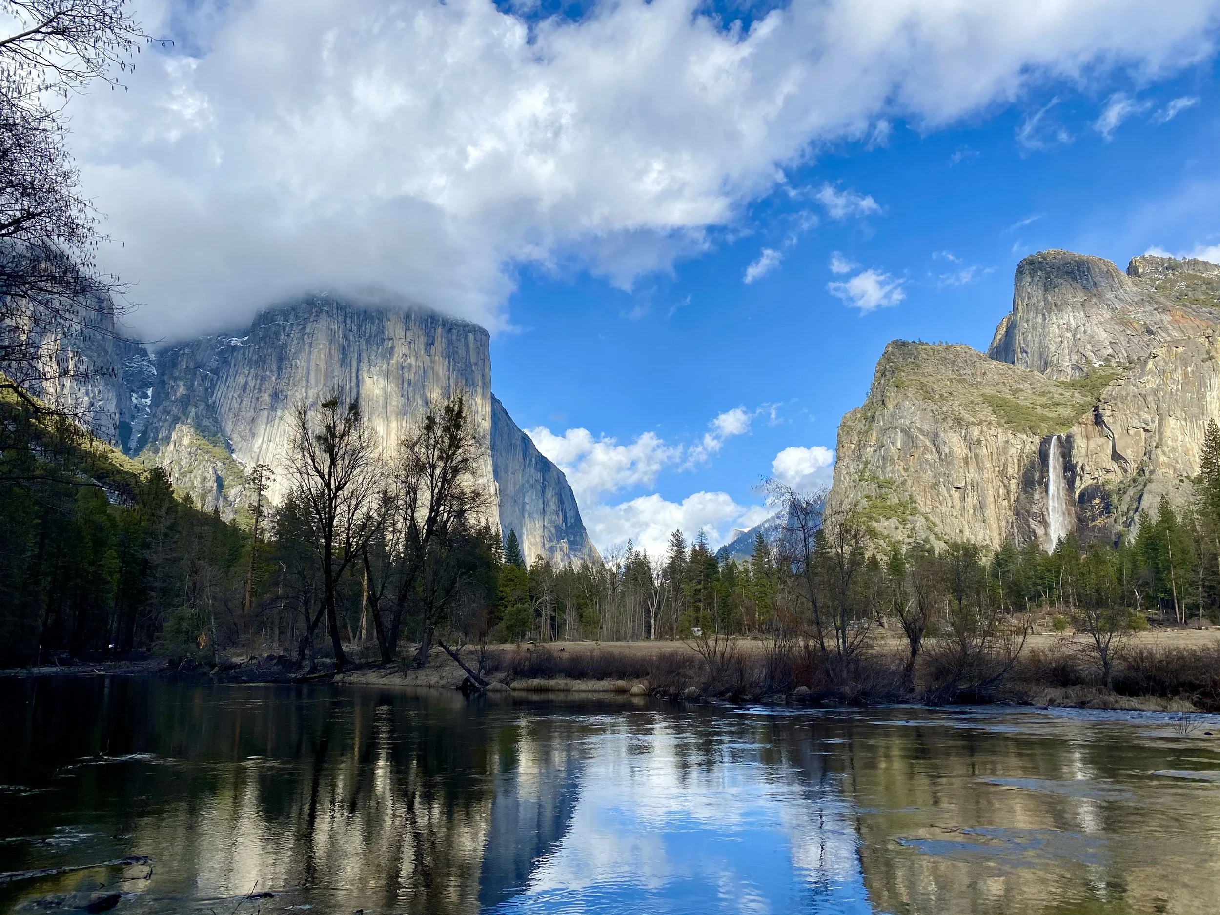Scenic view of a river surrounded by trees with tall granite cliffs and waterfalls, under a partly cloudy sky.