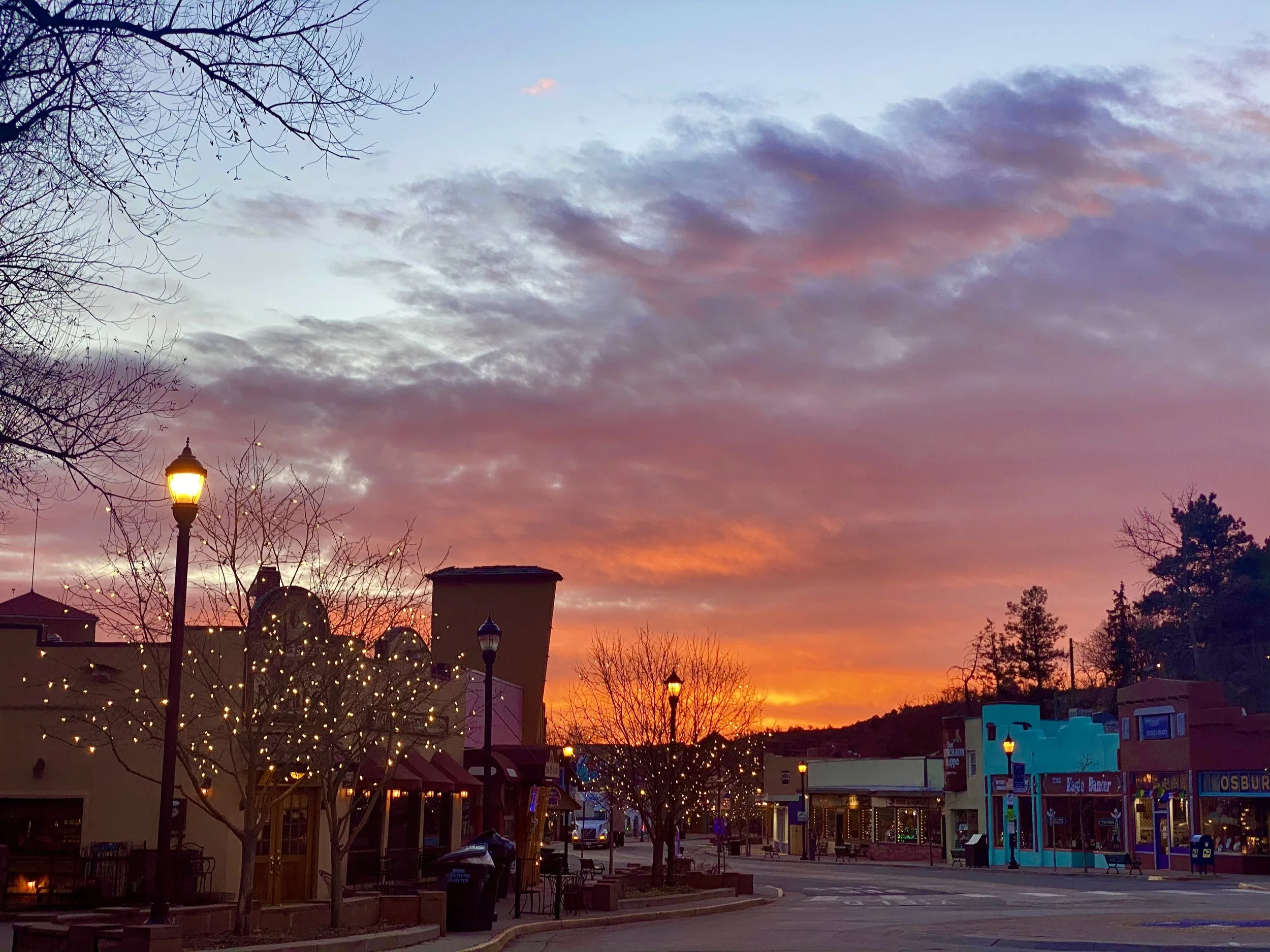 A small town plaza at sunset with trees, storefronts, lamp posts, and string lights, under a colorful sky with orange, pink, purple, and blue clouds.