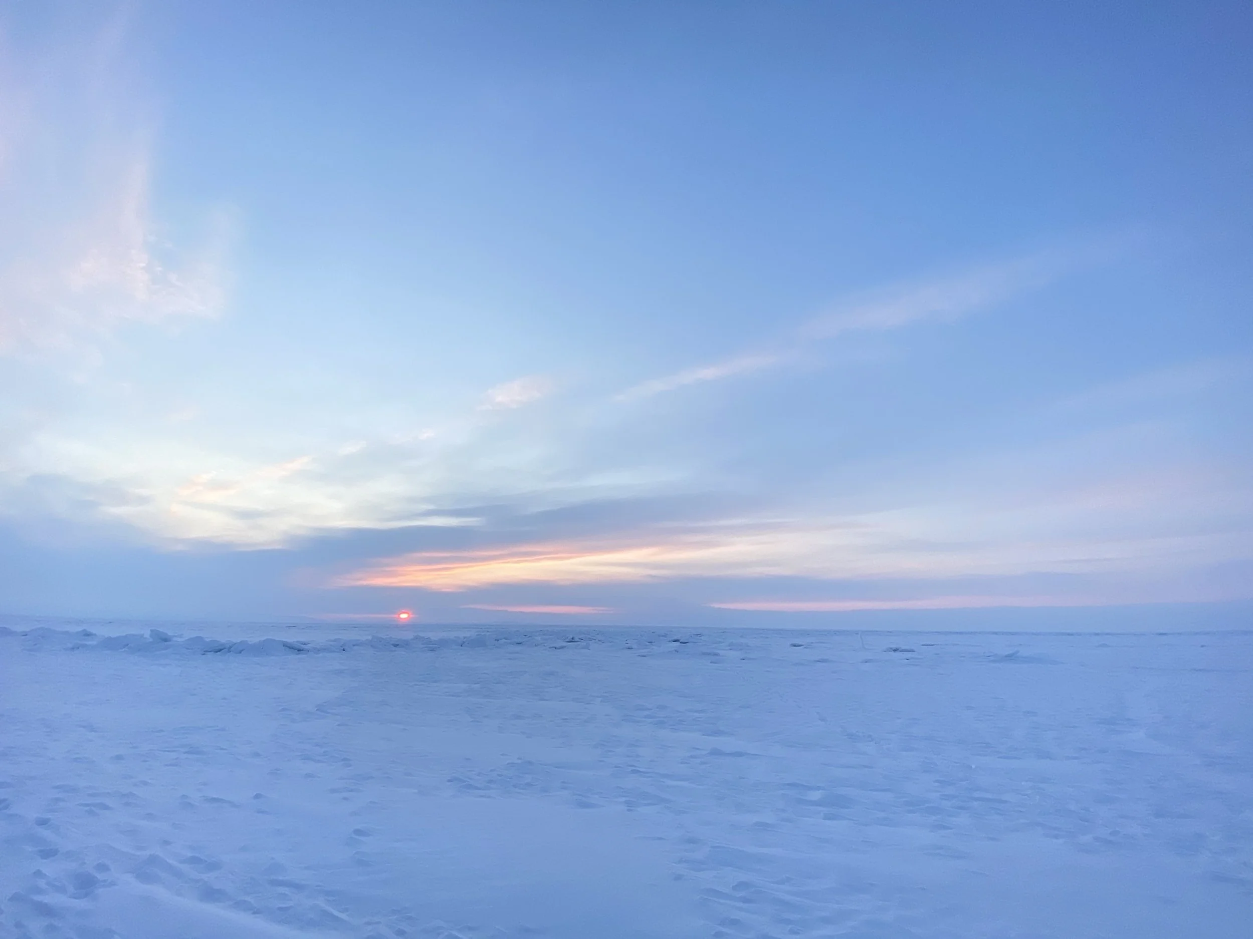 A snowy landscape during sunset with a mostly clear sky, some clouds, and the sun near the horizon.
