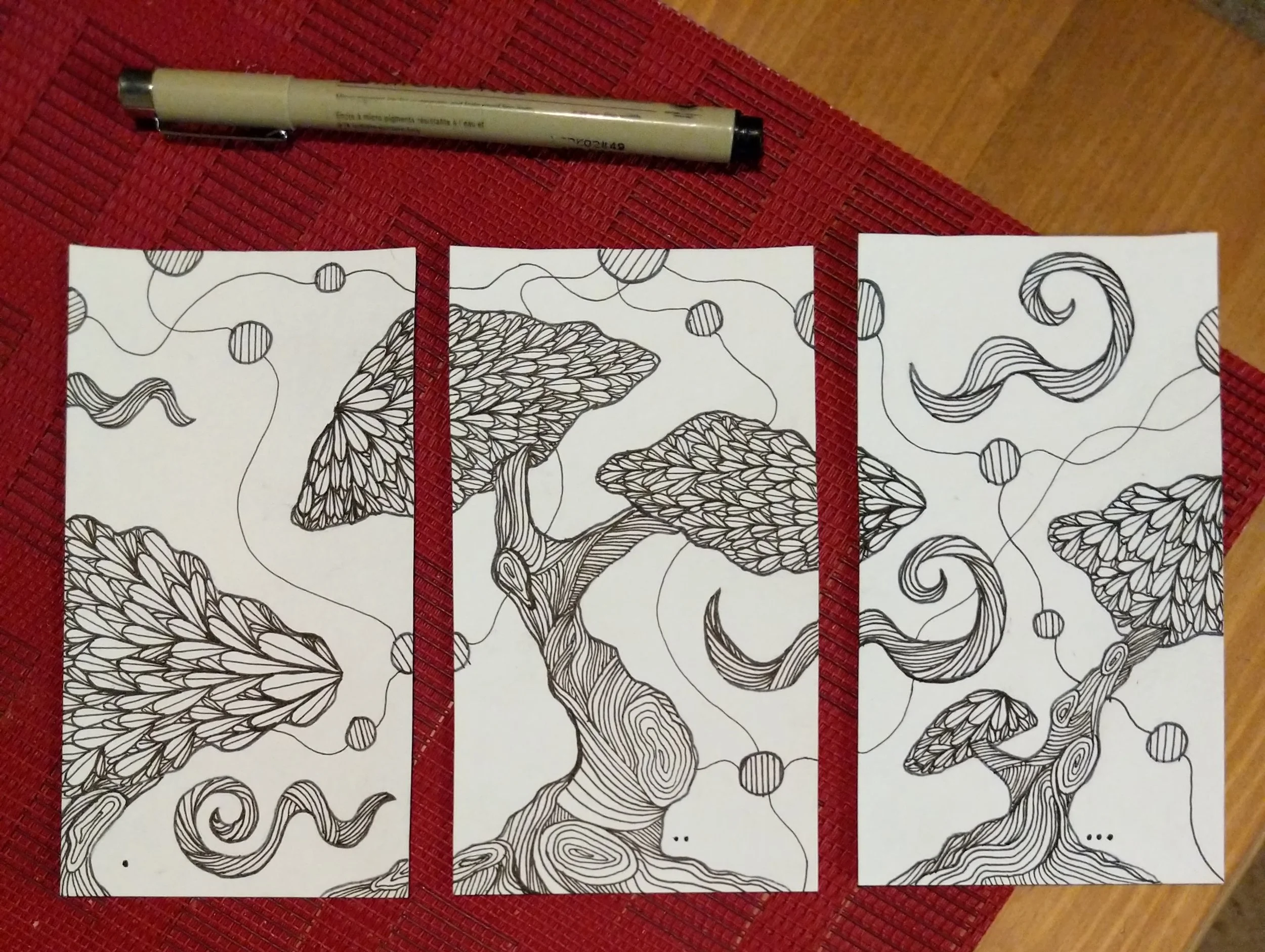 Three black and white abstract line drawings of trees with textured leaves and swirling branches, placed side by side on white paper, with a black pen above them, on a red fabric surface.