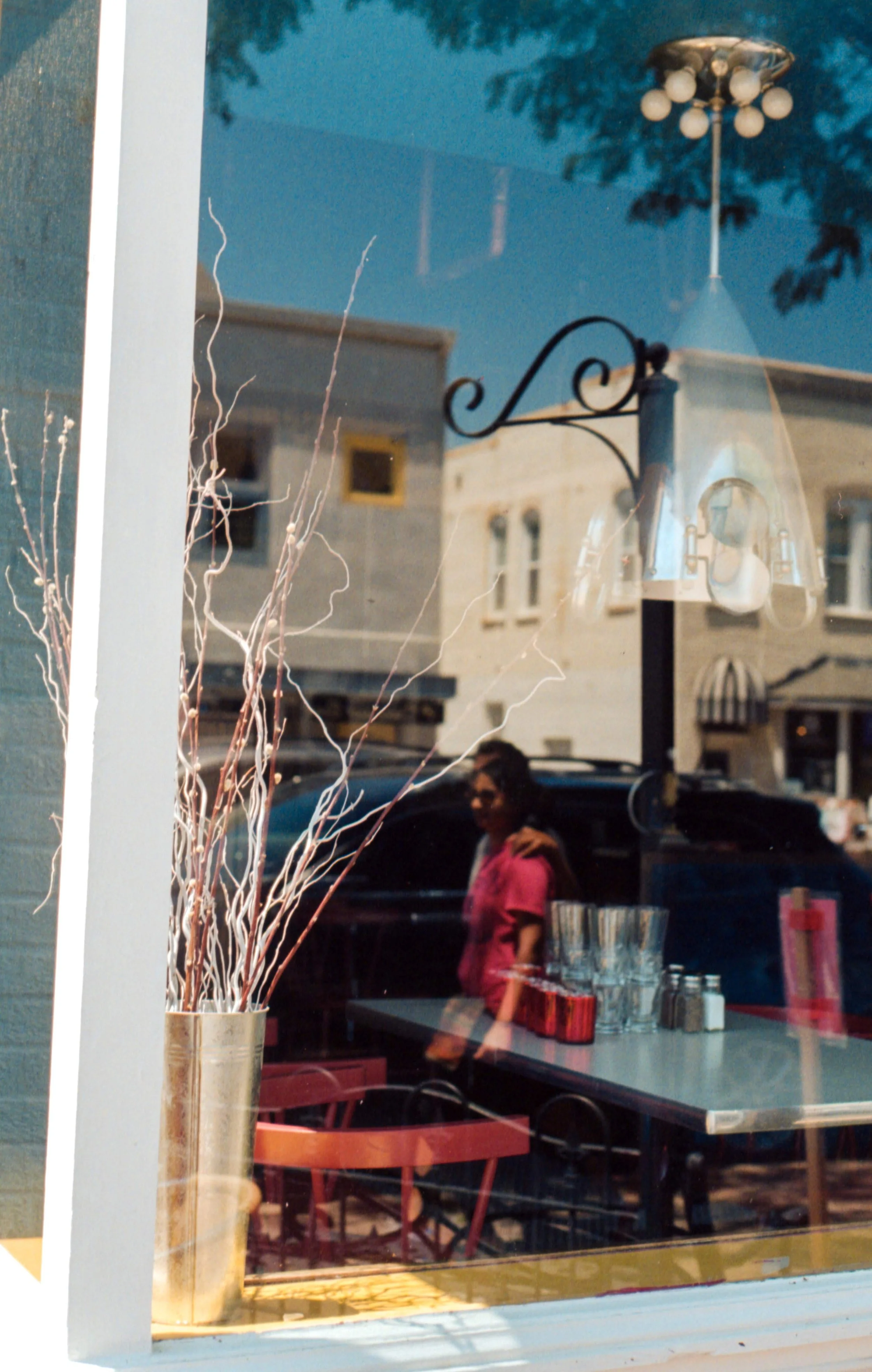 Reflected street scene seen through a window with a person inside a cafe in the background, chairs and tables inside, and buildings outside with a hanging light fixture.