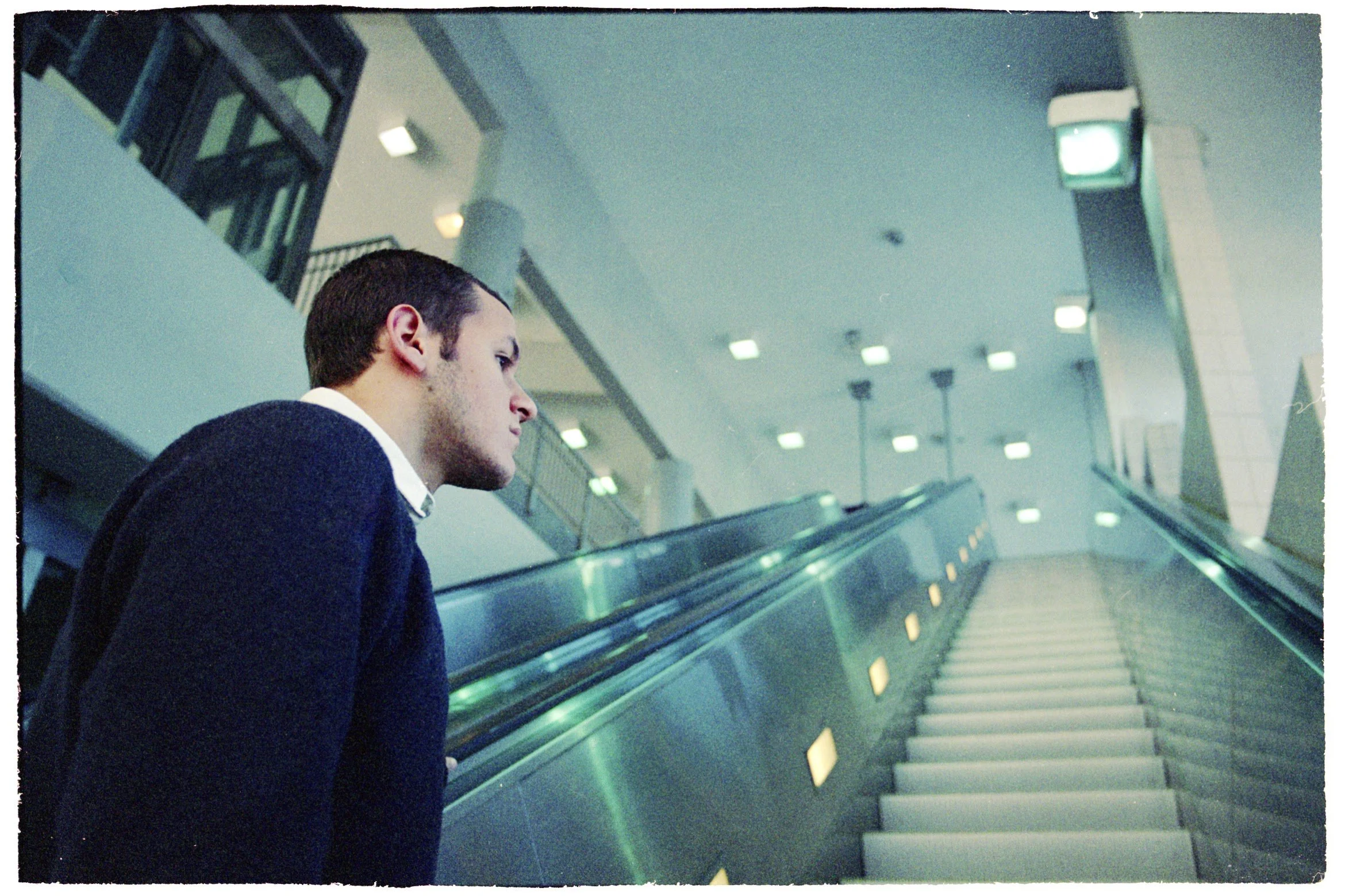 A young man dressed in a dark sweater and white shirt looking at an escalator in a modern, well-lit building.