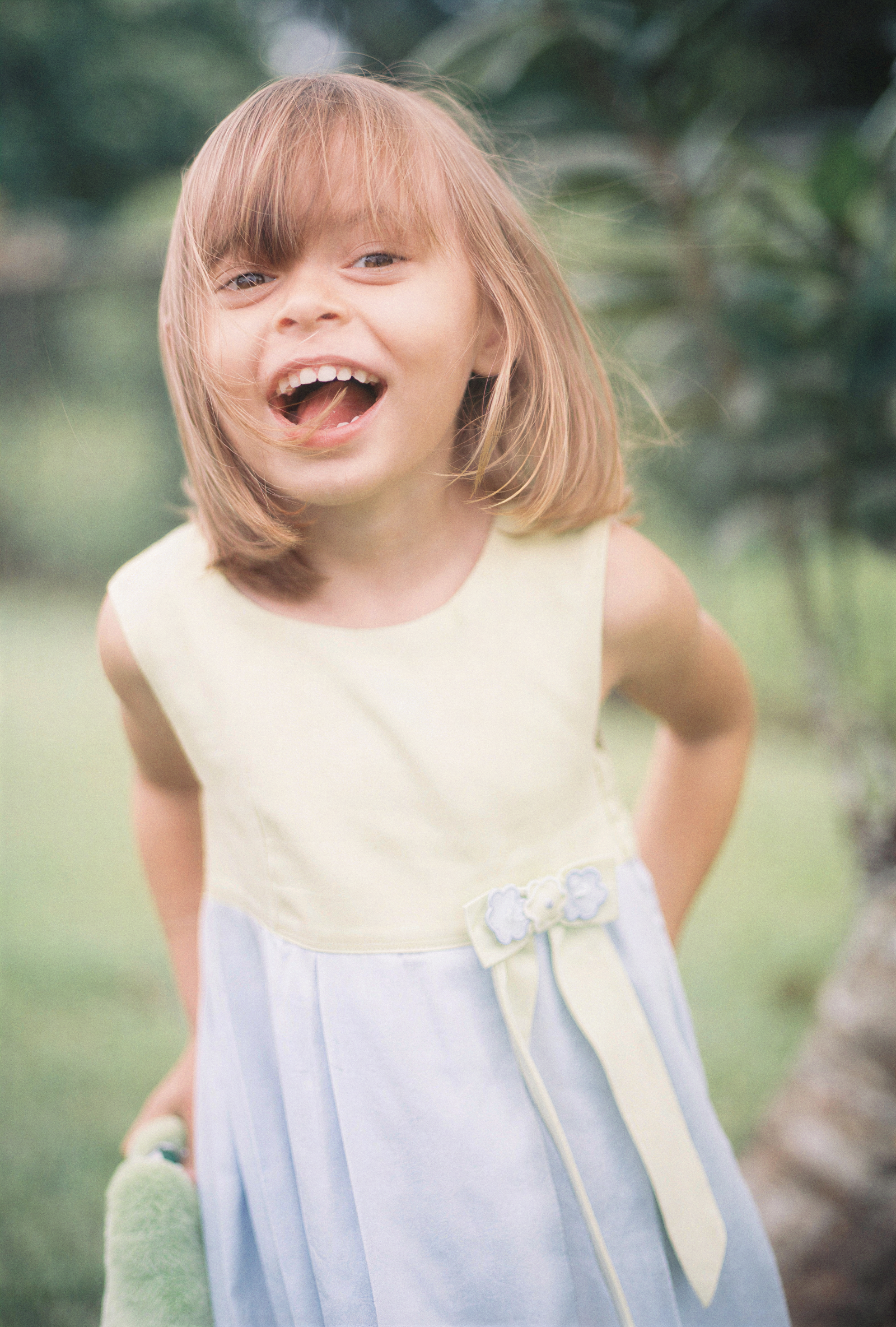 A young girl with shoulder-length light brown hair smiling outdoors in a garden, wearing a sleeveless yellow and gray dress with a flower and bow detail.