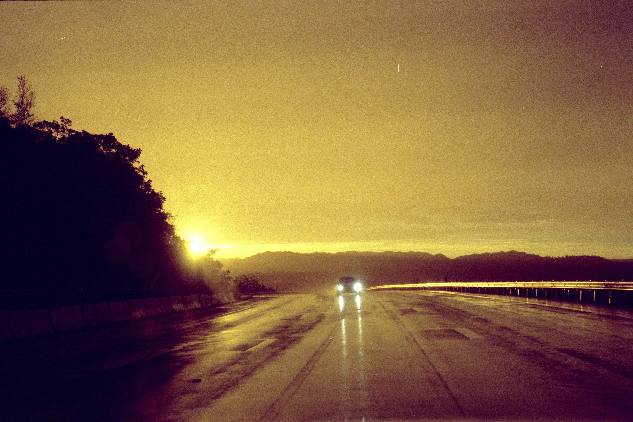 A car driving on a wet road at night, with headlights on, reflecting on the wet surface. The sky is yellowish with clouds and visible stars, and the silhouette of trees and distant hills are on either side of the road.