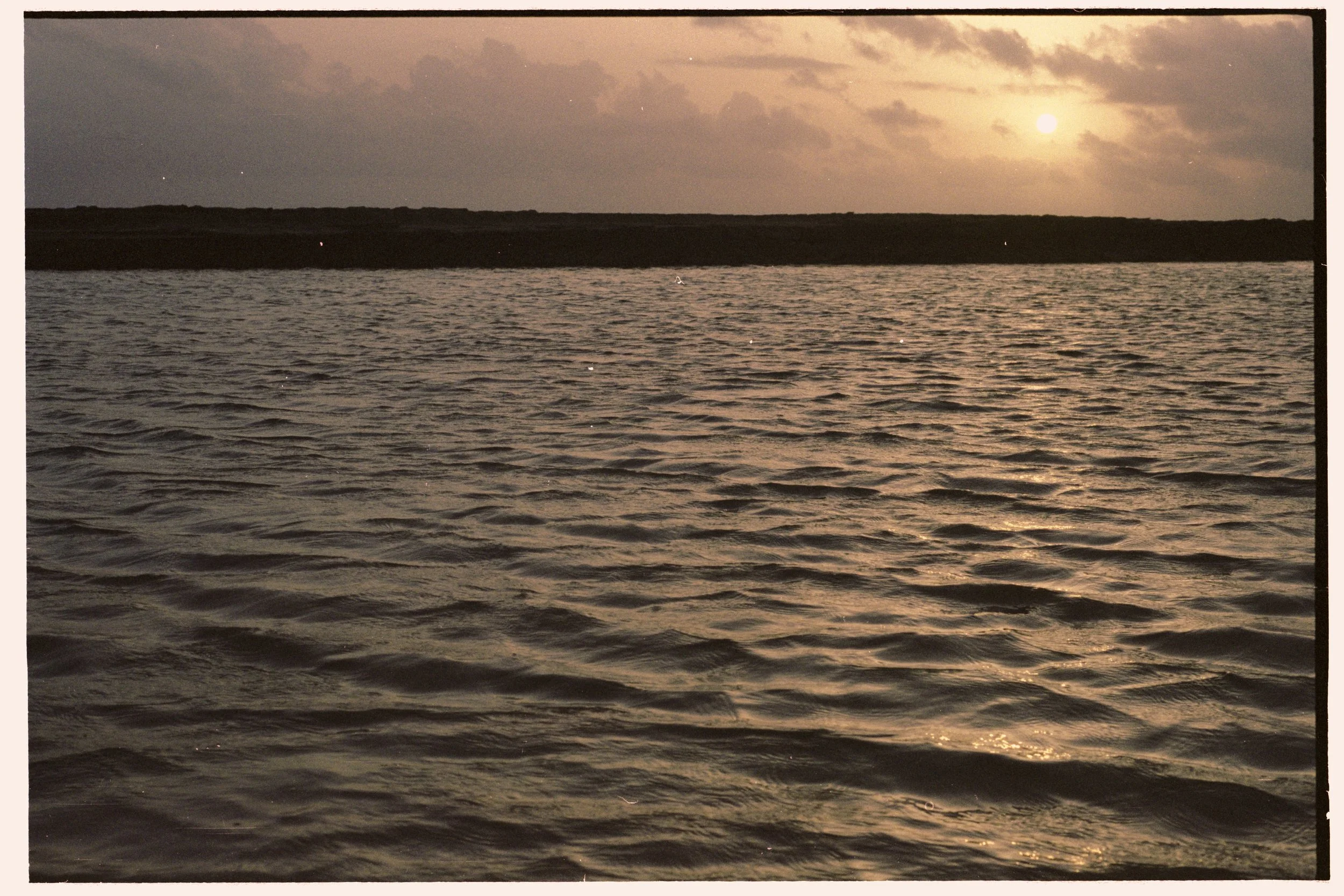 A body of water at sunset with the sun partially obscured by clouds and a dark horizon line in the background.