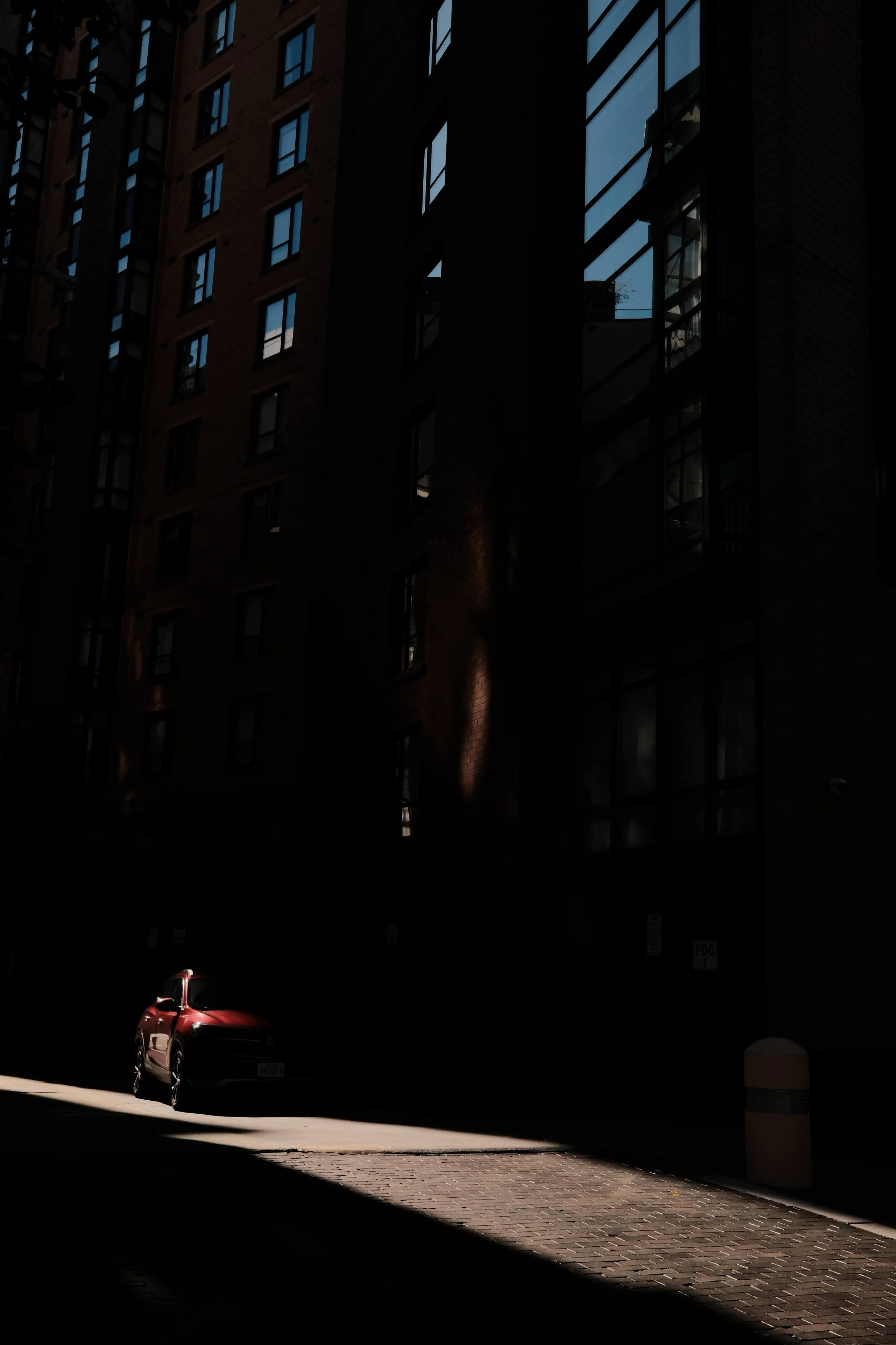 A red car parked on a brick-paved street in a shadowed alleyway with tall apartment buildings and large glass windows.
