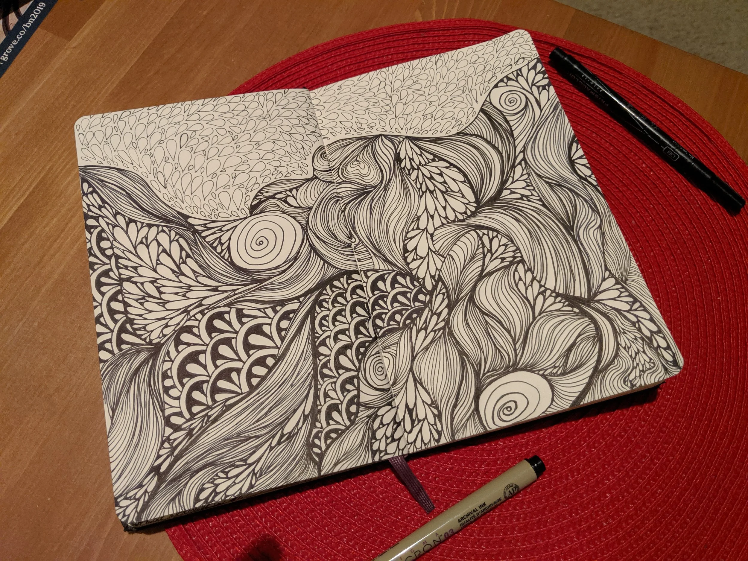 Open black and white abstract doodle art journal with intricate patterns and swirls, placed on a red textured placemat on a wooden table, with a black marker pen on the top right and a gray marker pen at the bottom.
