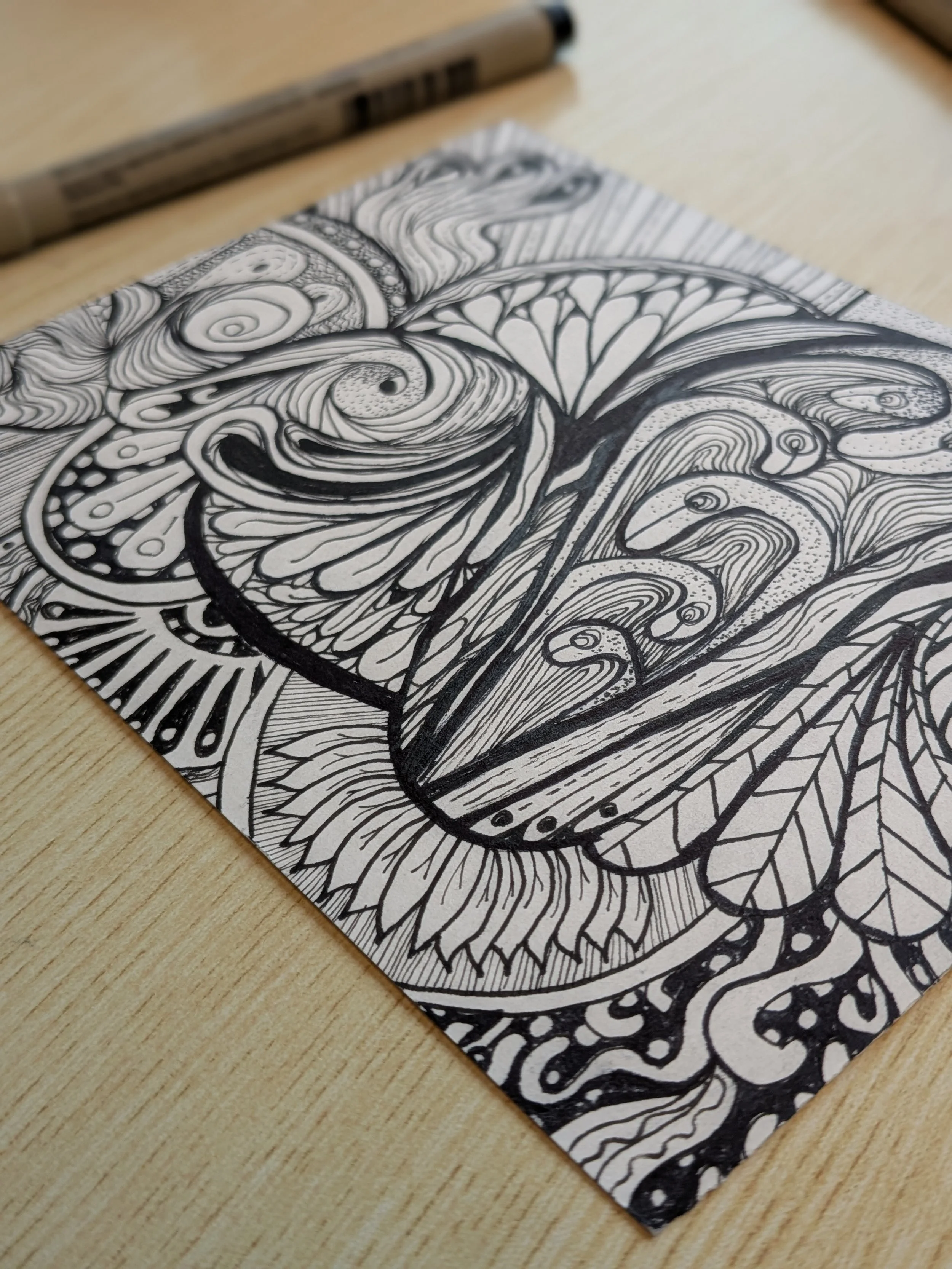 Black and white doodle art with intricate patterns and swirling designs on a sheet of paper, with a pen nearby on a wooden surface.