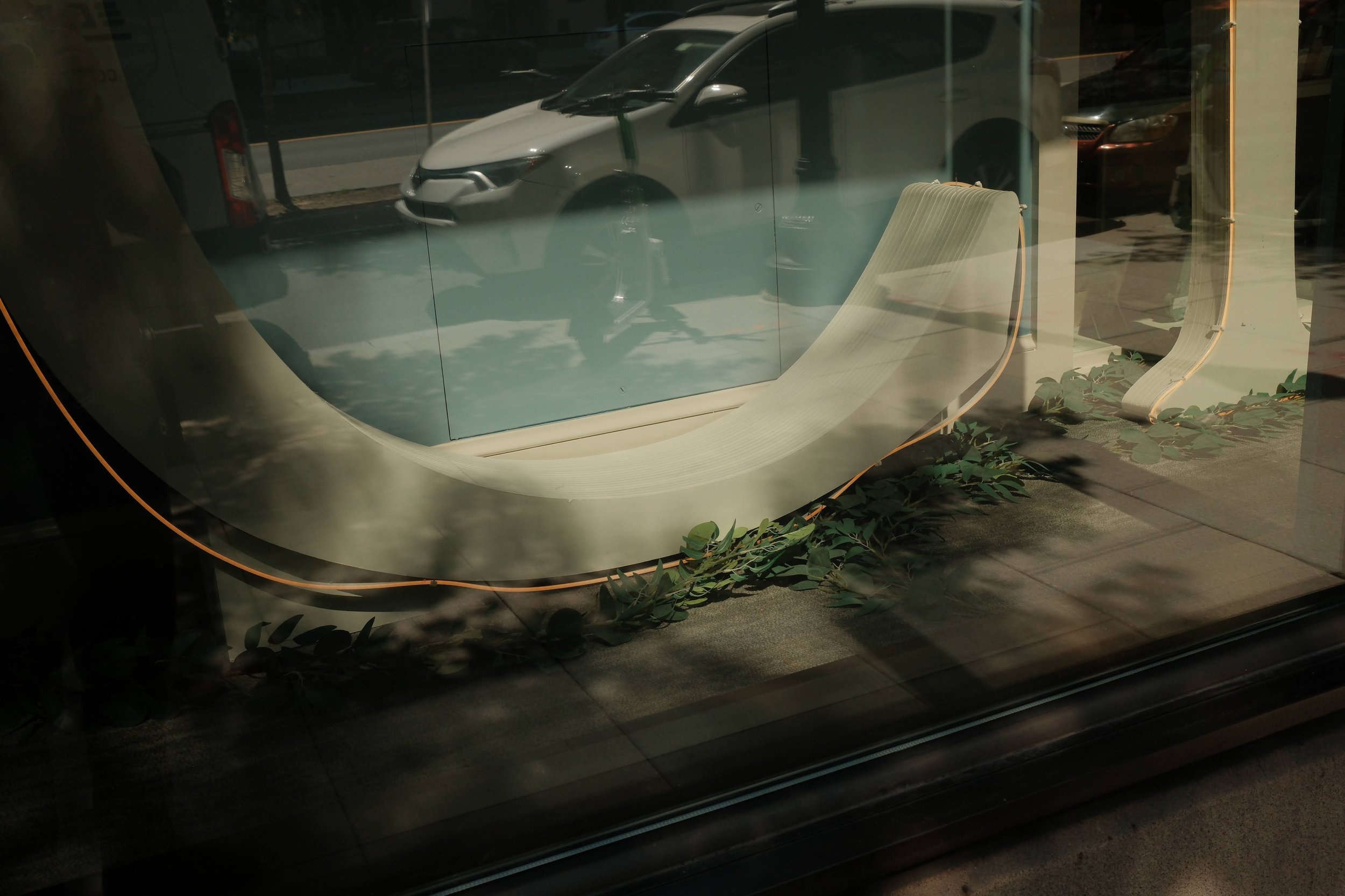 Modern cafe window display with a minimalist white bench and chairs, visible through glass, with reflections of parked cars and street scene outside.