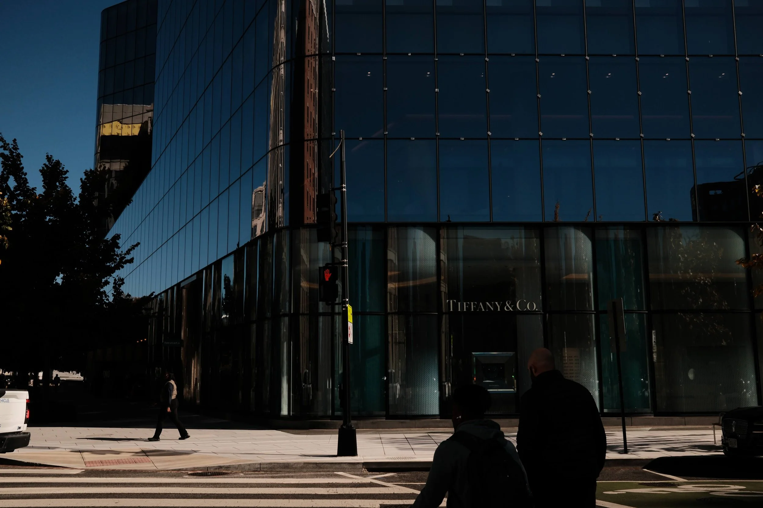 Exterior of a modern glass building with the Tiffany & Co. logo, people crossing the street, and a traffic light.