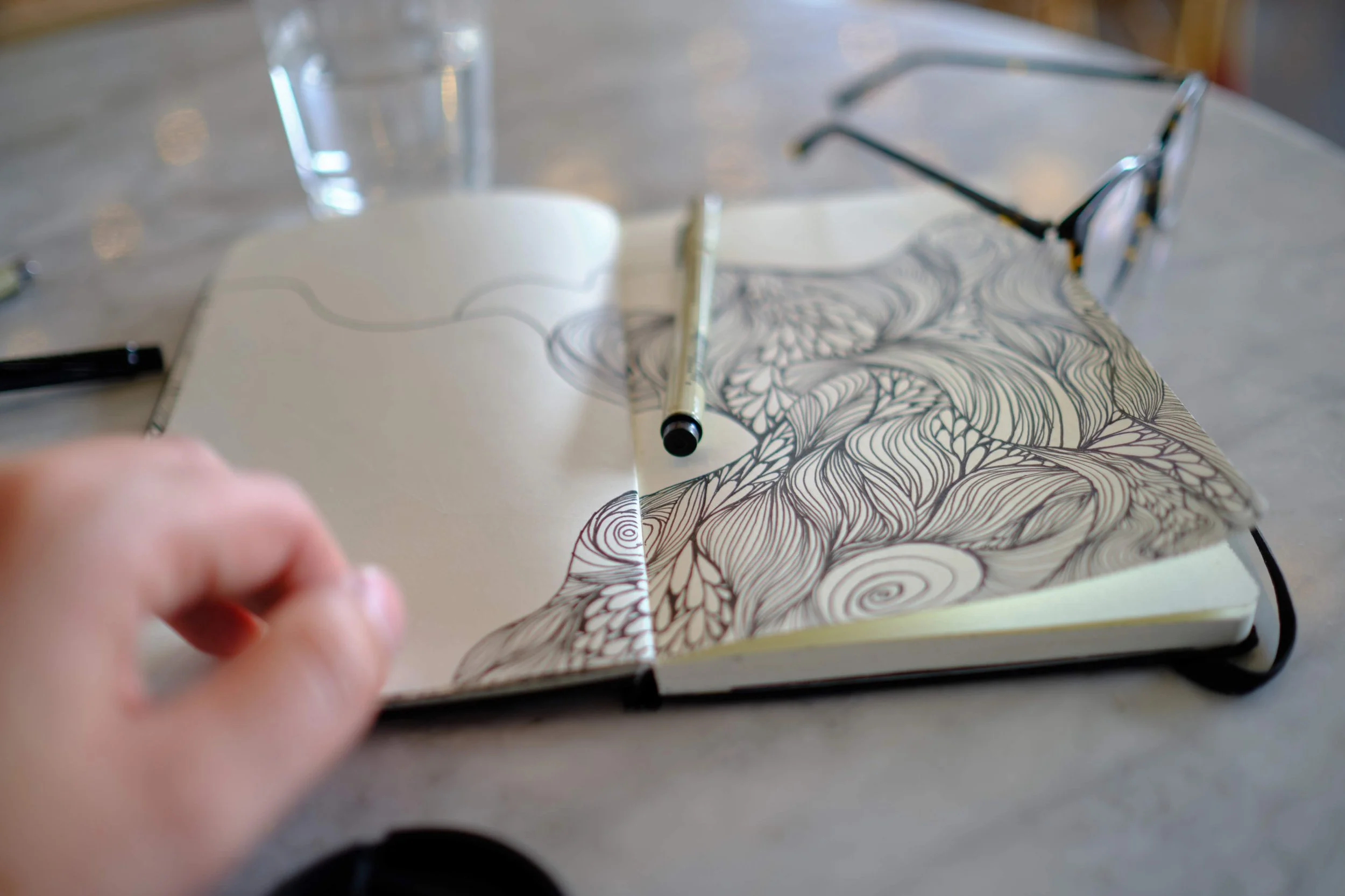 Open sketchbook with intricate black and white line art on pages, a fine-tipped pen resting on the book, a glass of water, and a pair of sunglasses on a light-colored table.