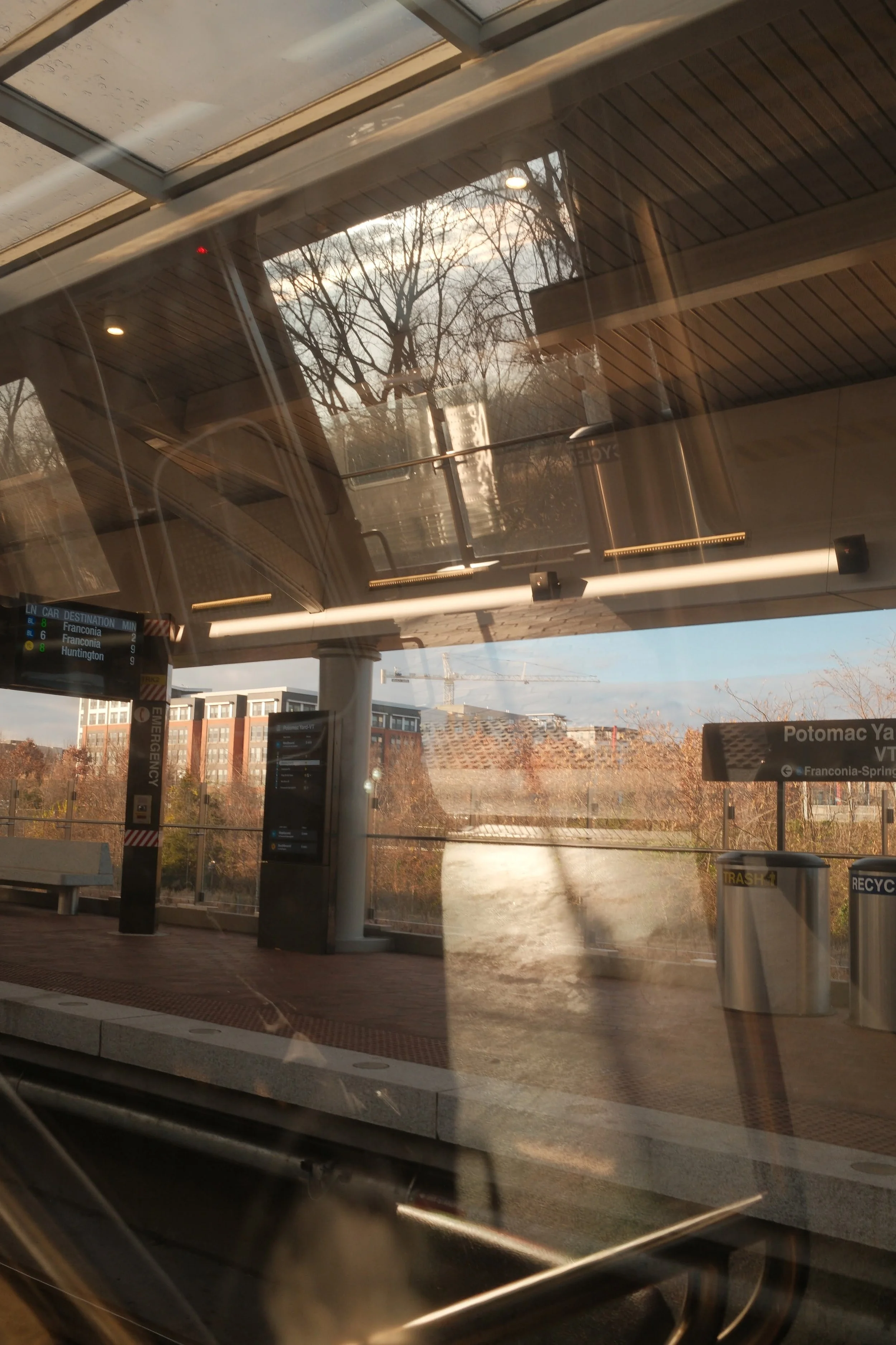 Reflections of trees and buildings on a train station platform with trash and recycling bins, a digital transit sign, and a glass shelter.