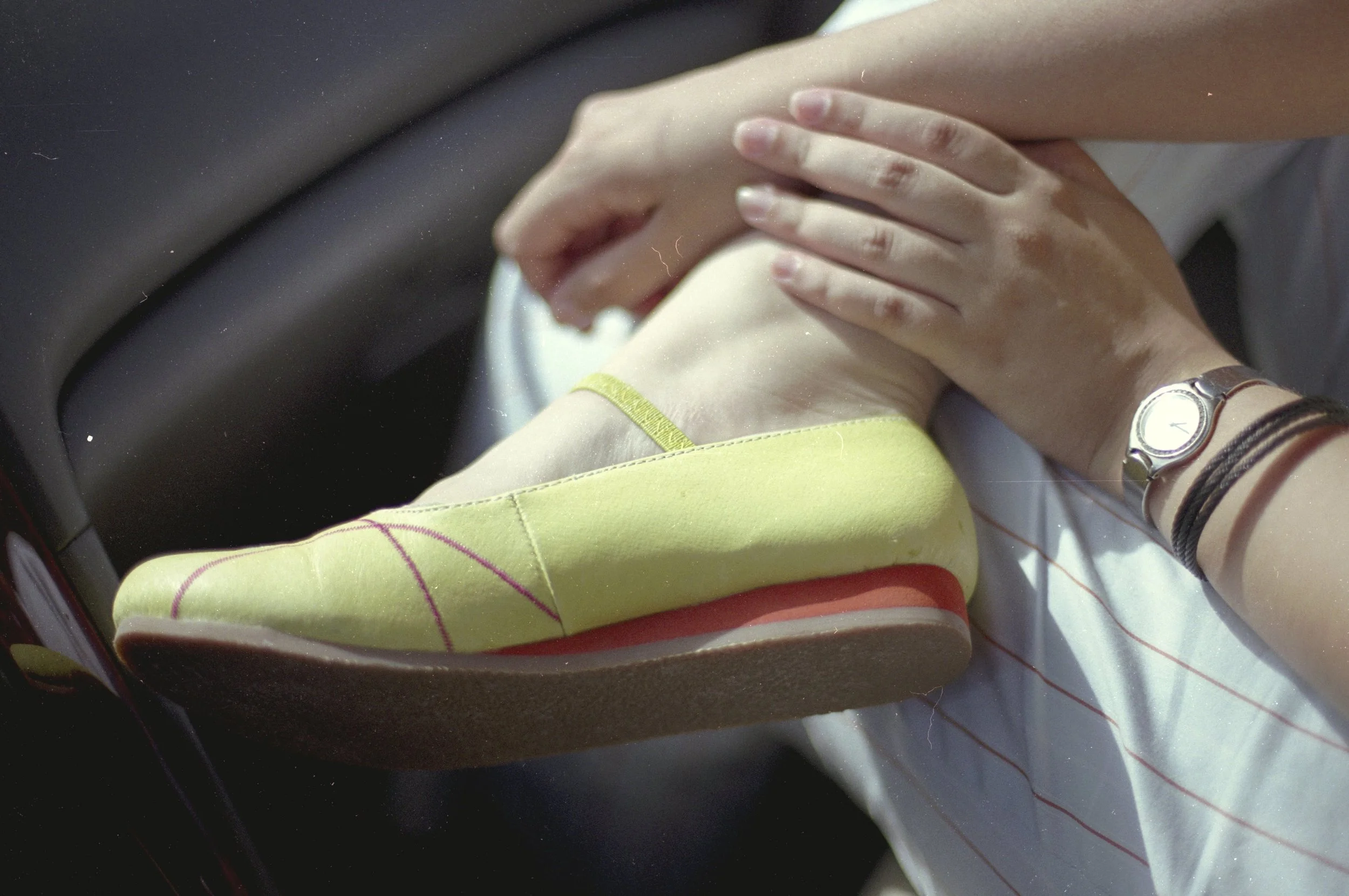 Close-up of a woman putting on a bright yellow slip-on shoe with red sole. She is wearing a silver watch and a black bracelet on her wrist, and a white shirt with thin red stripes. The background shows part of a car interior.