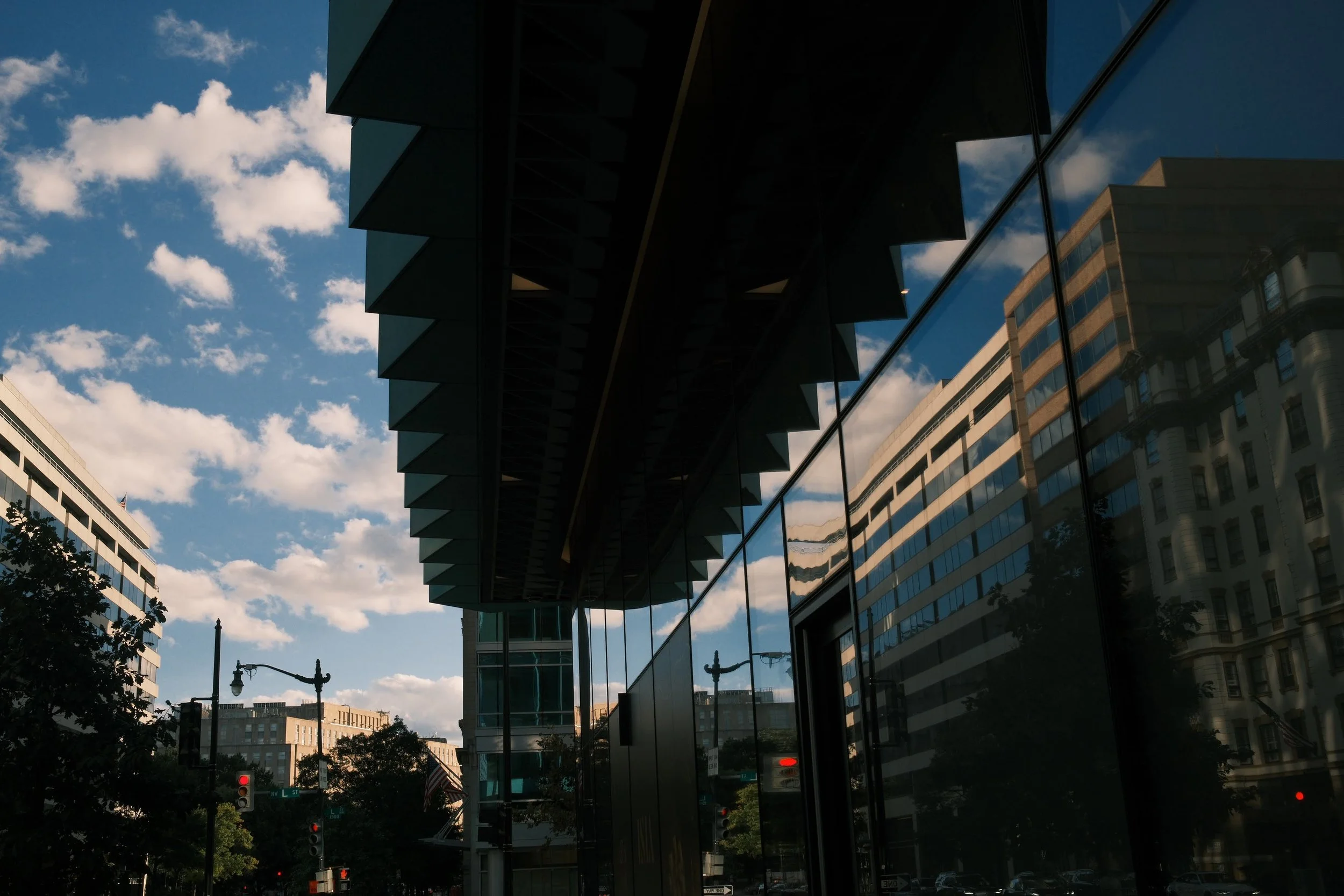 City street scene with modern glass buildings, trees, traffic lights, and a partly cloudy sky