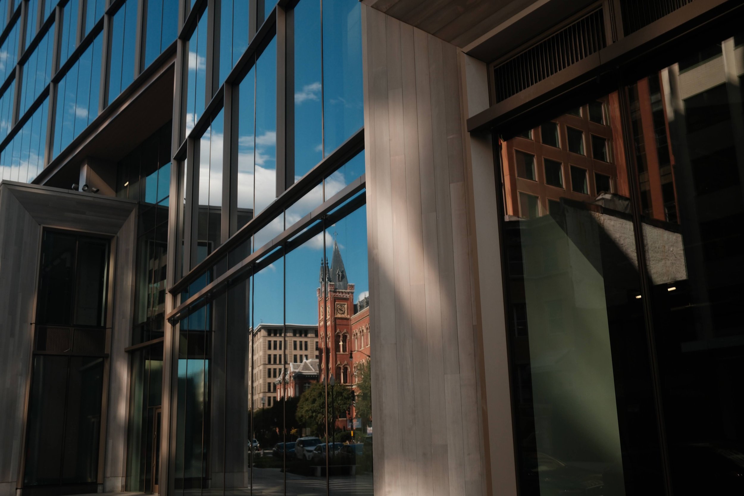 Reflection of historic church with tall clock tower and spire in the glass windows of a modern building with wooden and glass facade.