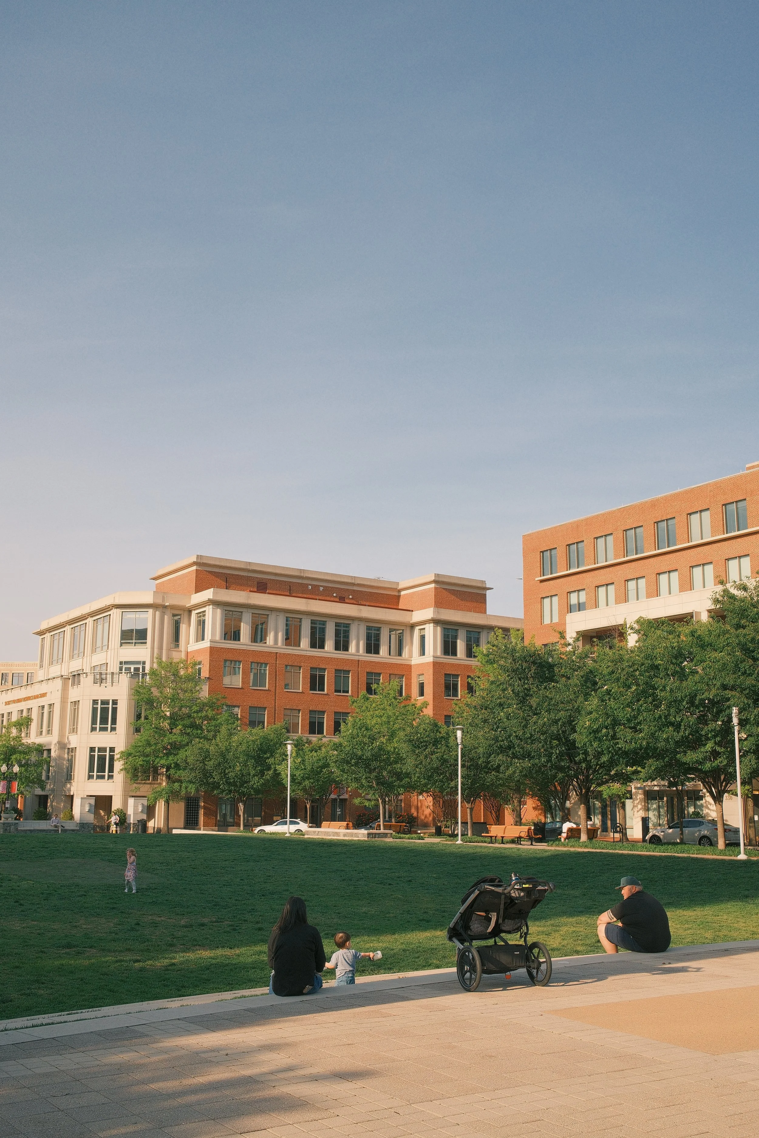 People sitting on a sidewalk near a grassy area with trees and modern buildings in the background, during daytime.