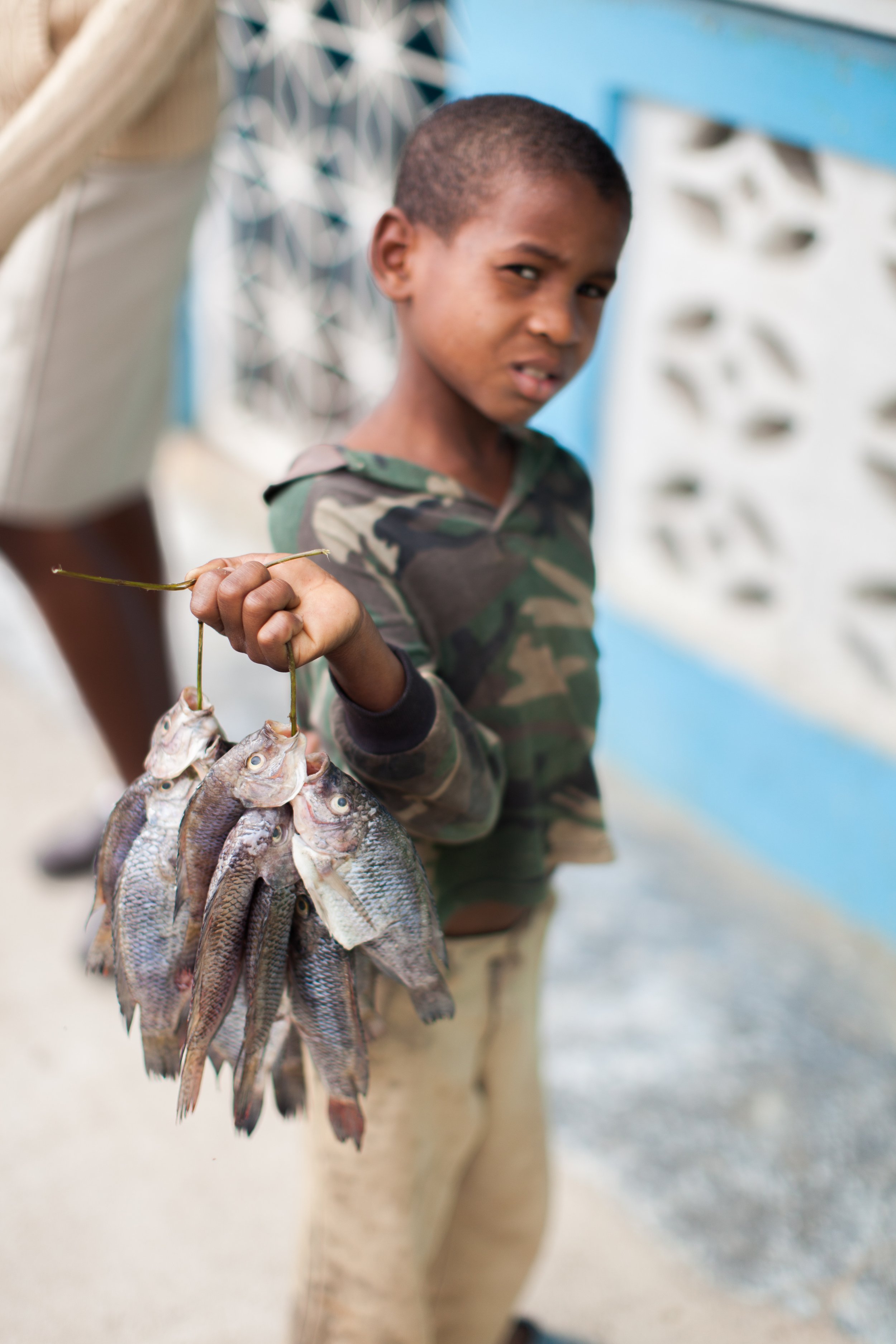 A young boy holding a string of freshly caught fish, standing outdoors near a wall with a geometric pattern.