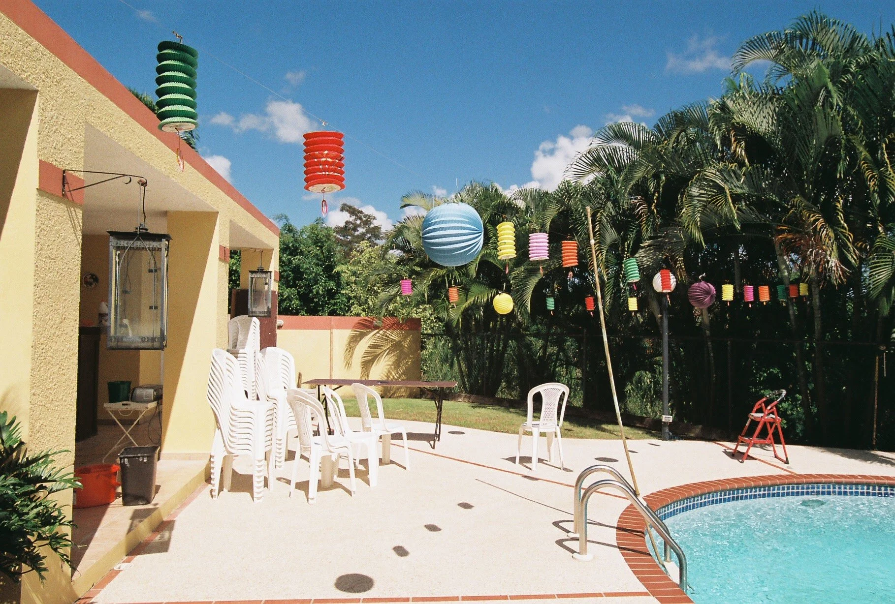 A backyard with a swimming pool, potted plants, and stacked white plastic chairs. Colorful lanterns hang from above, with palm trees and a clear blue sky in the background.