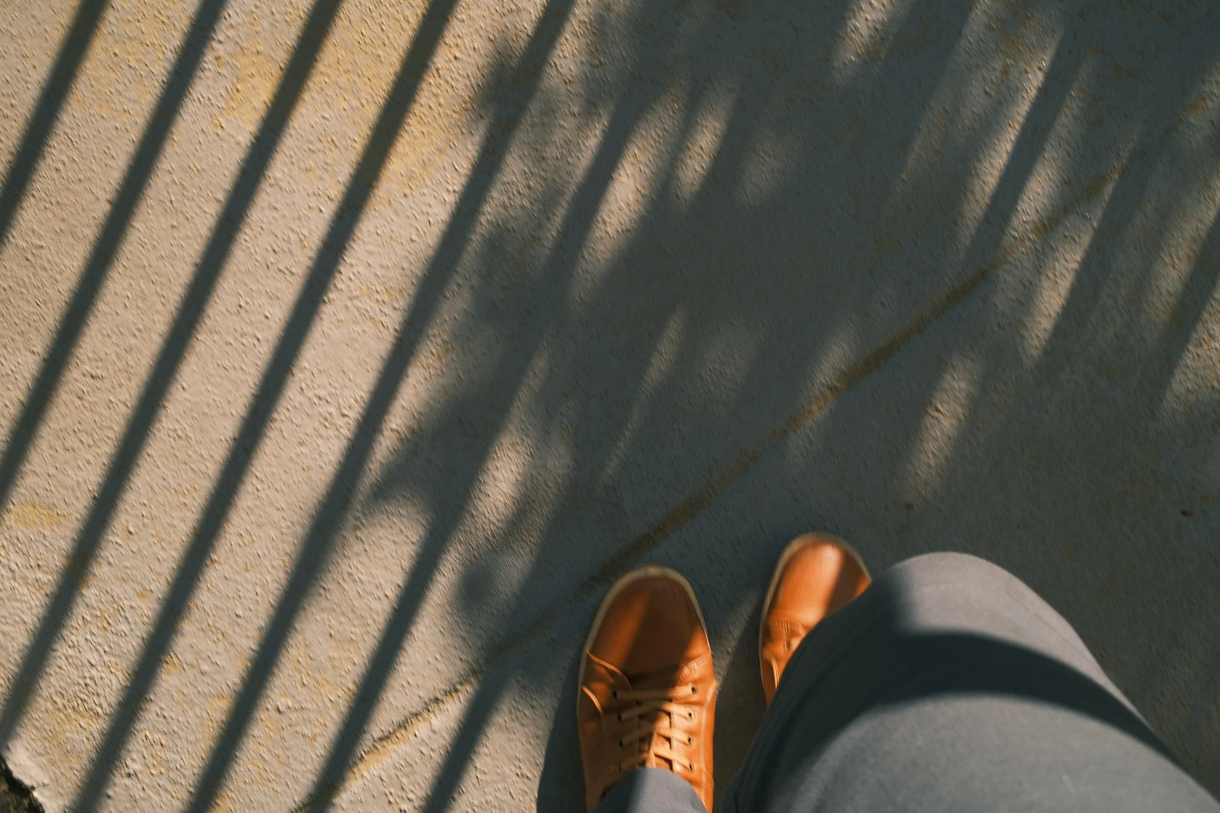 A person standing on a sandy surface, wearing brown shoes and grey shorts or pants, with long shadows cast by a railing or fence outside.