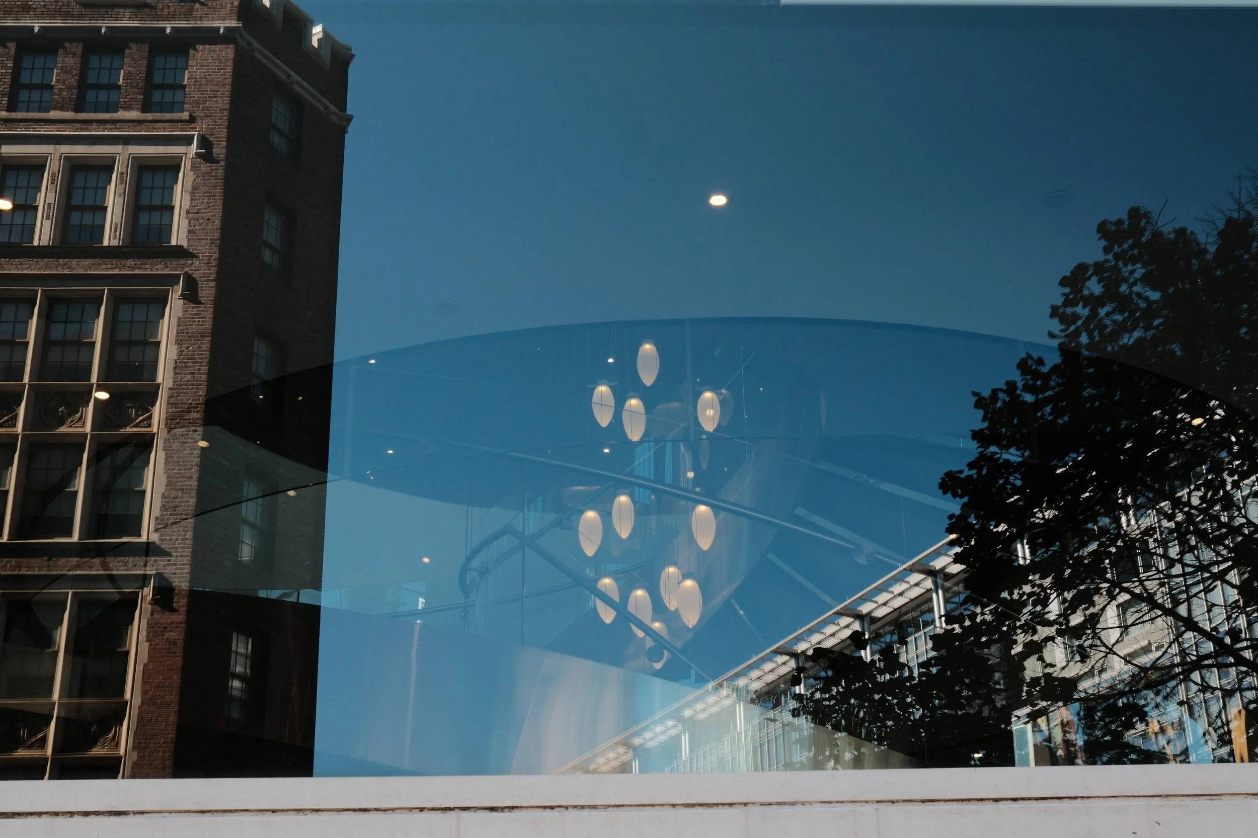 View through a glass window showing reflections of a building, tree, and indoor lighting fixtures against the blue sky.