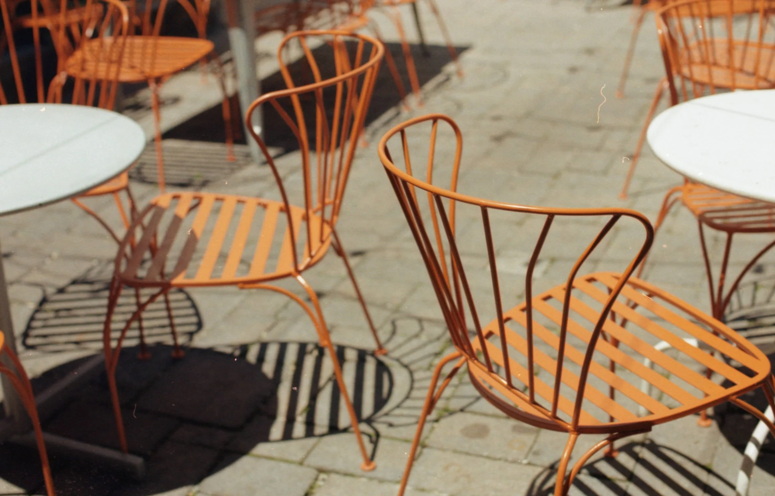 Several orange metal outdoor chairs and white round tables on a cobblestone patio, with shadows cast by the chairs