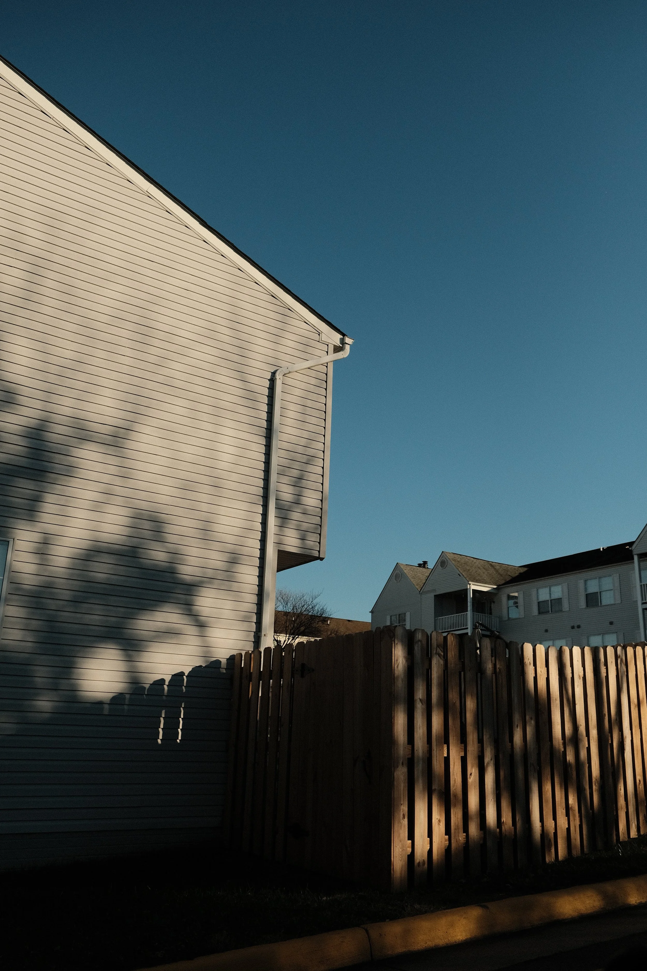 A side view of a beige house with horizontal siding and a white gutter, shadow of a tree cast on the house, with a wooden fence in the foreground and other houses visible in the background under a clear blue sky.
