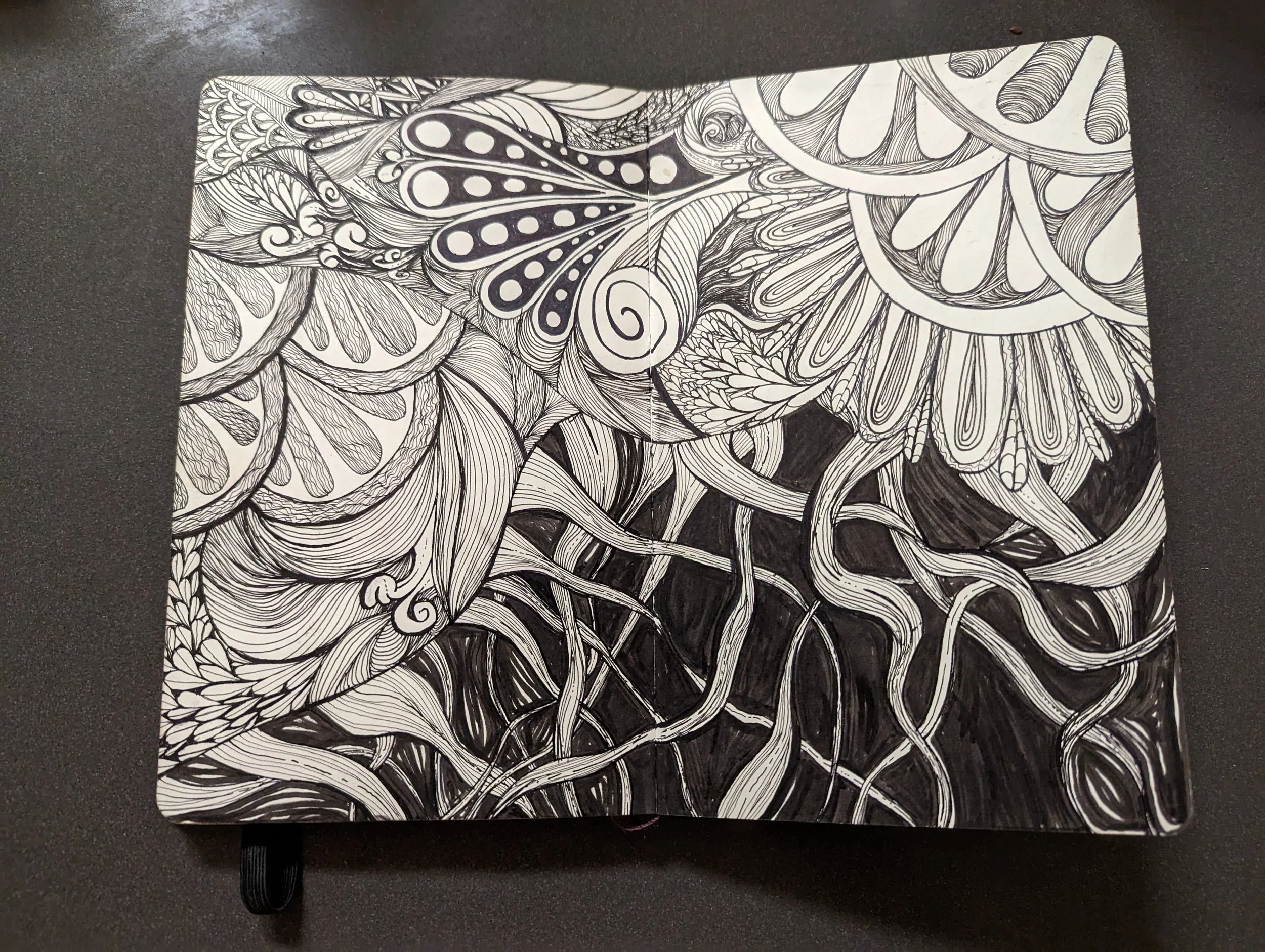 A black and white abstract doodle drawing with intricate patterns, swirls, lines, and shapes, partly filling a notebook on a dark surface.