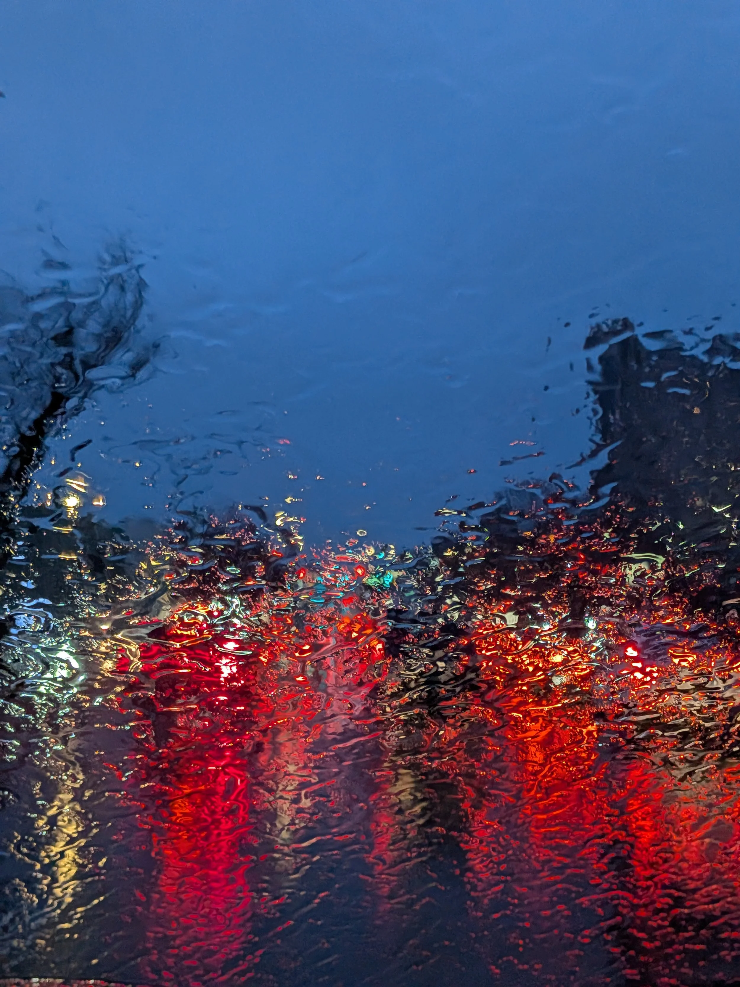 Rainy weather seen through a car windshield with blurred red, yellow, and blue lights reflecting off the wet surface.